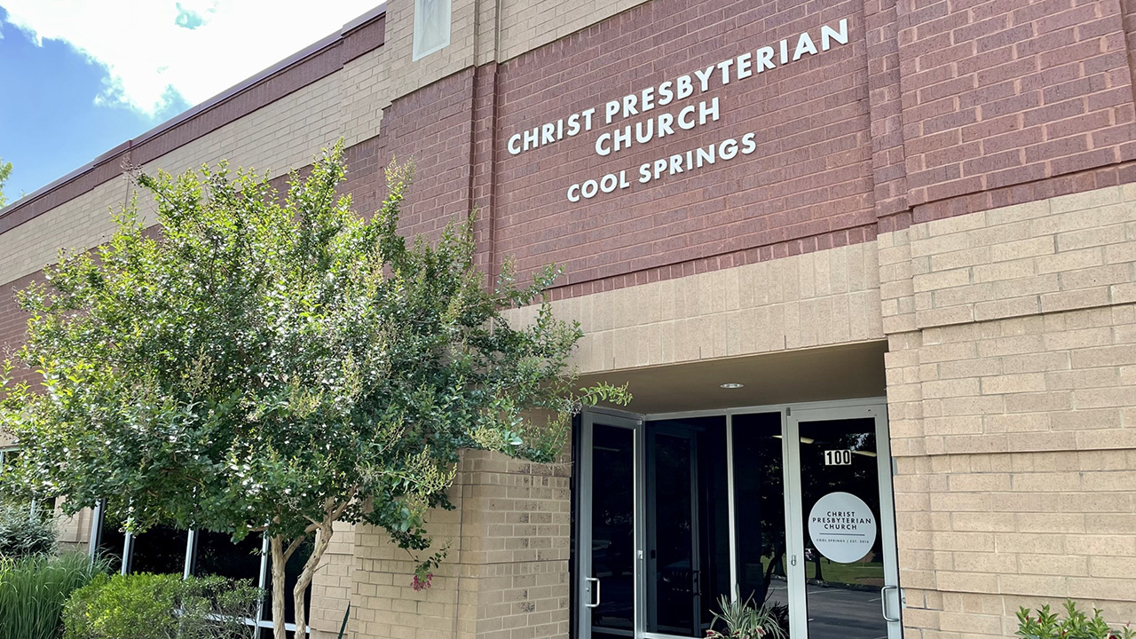 Exterior of Christ Presbyterian Church Cool Springs, a brown brick building with large windows and a glass entrance door, surrounded by green trees and shrubs under a partly cloudy sky.