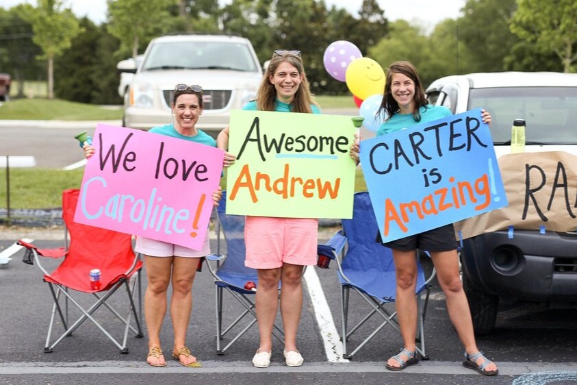 Three women stand in a parking lot holding colorful signs that read We love Caroline!, Awesome Andrew, and CARTER is Amazing! They smile by parked cars and balloons, celebrating all ministries. Learn more about this joyful gathering!.