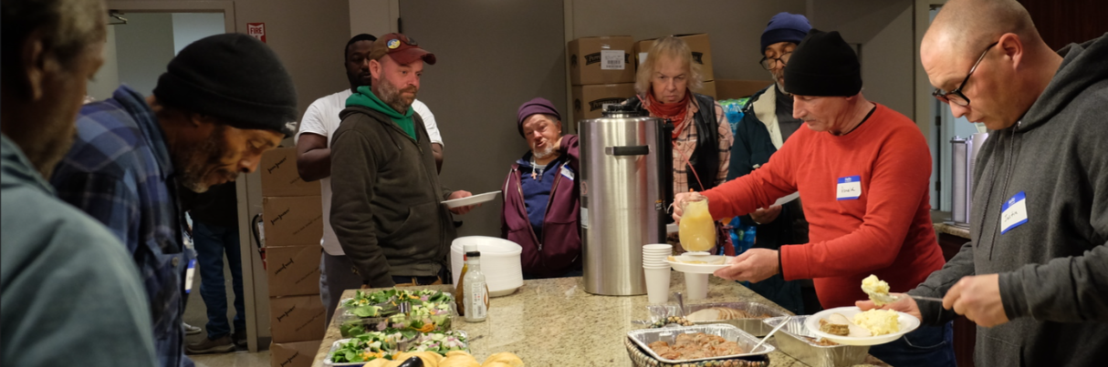 A group of people, some wearing hats and coats, gather around a counter serving food and drinks. Plates of salad, bread, and hot dishes are on the counter. Two people are serving themselves from the trays.