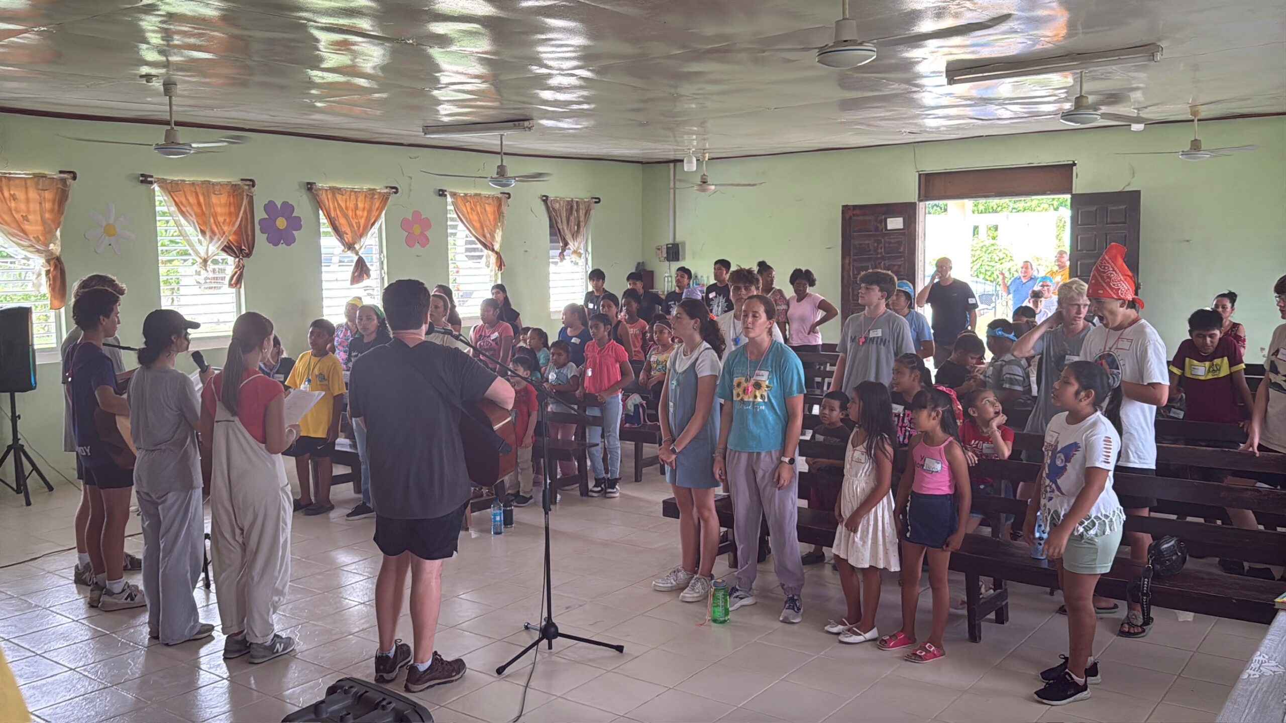 A group of people, including adults and children, stand inside a brightly lit room with benches, singing together. Some hold song sheets while a person plays guitar. Windows with orange curtains and flower decorations are visible.
