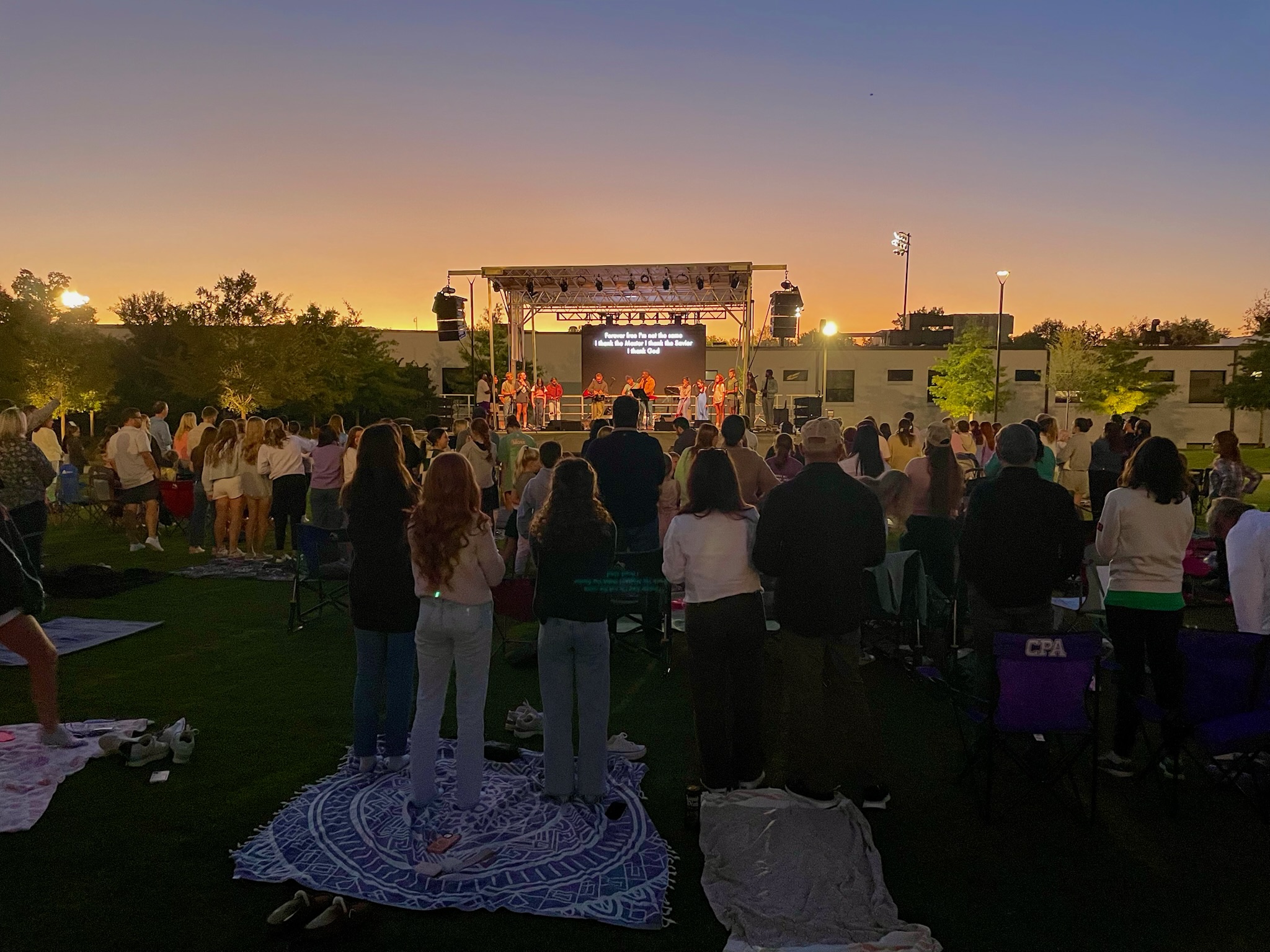 A crowd of people stands and watches a live outdoor concert at sunset, facing a brightly lit stage. Some sit on blankets and lawn chairs on a grassy field, while buildings and trees are visible in the background.