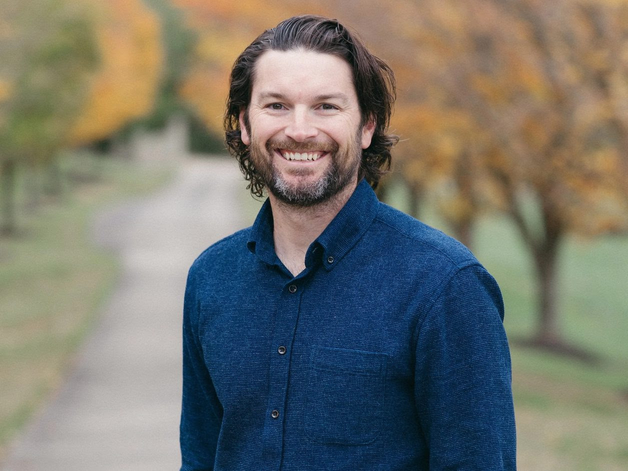 A man with medium-length brown hair and a beard smiles while standing outdoors on a pathway, with autumn trees and green grass in the background. He is wearing a blue button-up shirt.
