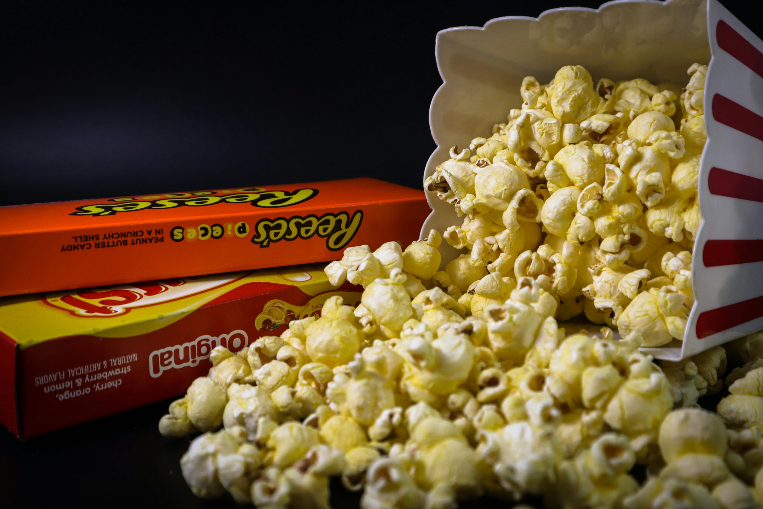 A striped popcorn container tipped over, spilling popcorn onto two boxes of candy: Reeses Pieces and Original Milk Duds, against a dark background.