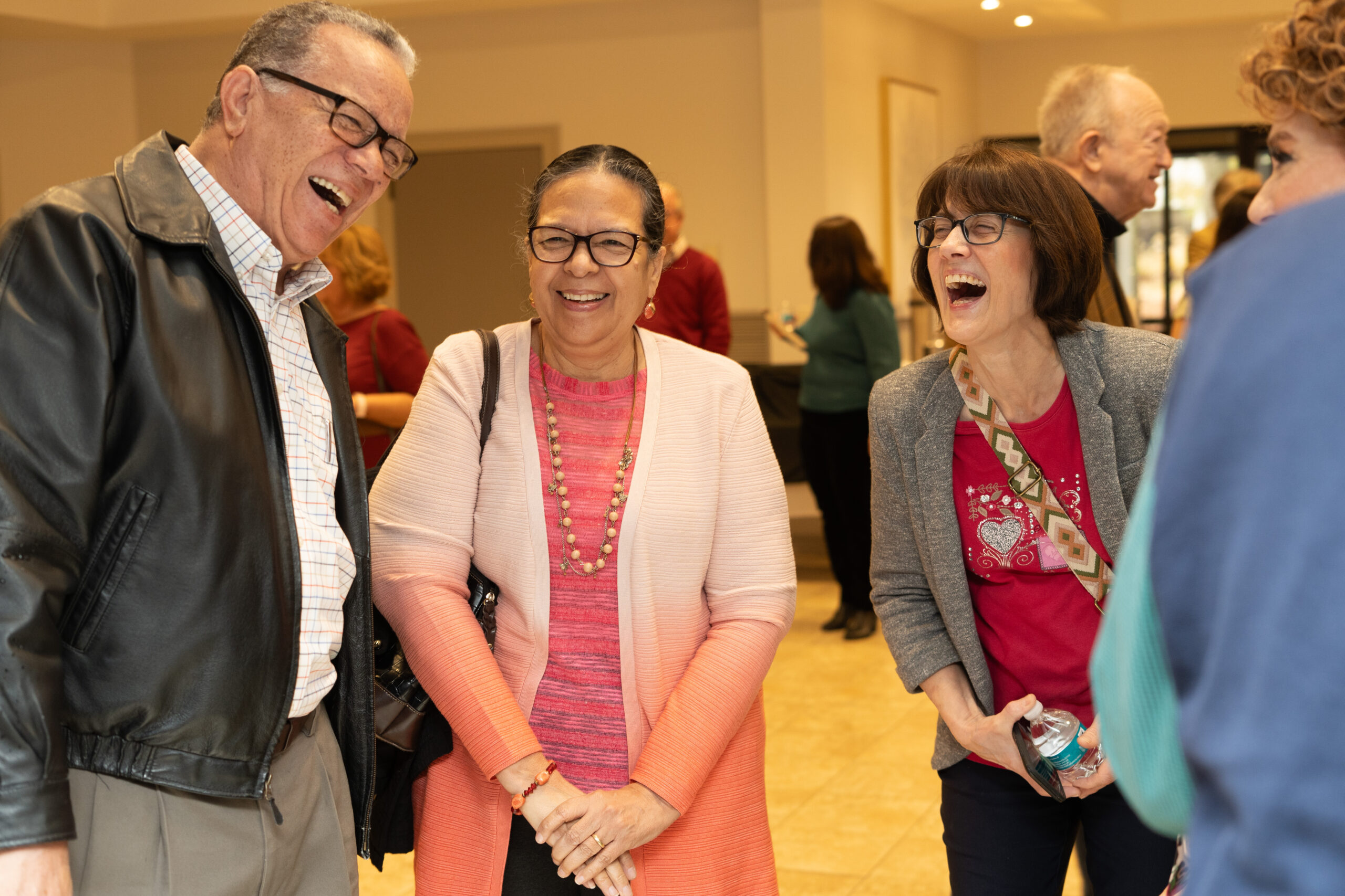 Three adults stand together indoors, laughing and smiling. The group appears to be enjoying a friendly conversation in a brightly lit room with other people chatting in the background.