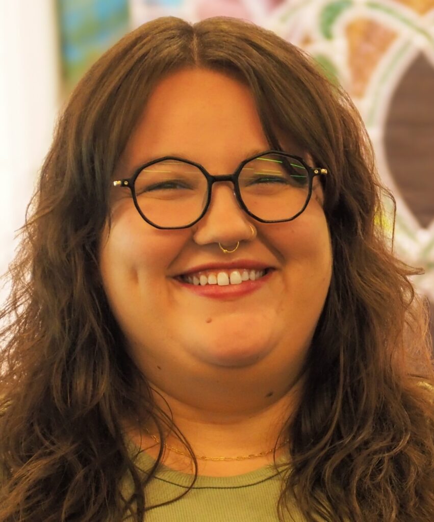 A person with long brown hair, glasses, and a septum ring smiles warmly at the camera. They are wearing a light green top and a gold necklace, with a blurred colorful background behind them.