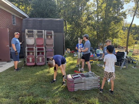 A group of people, including children and adults, are stacking and organizing folding chairs outside near a brick building and shed on a sunny day. Trees and more stacked chairs are visible in the background.