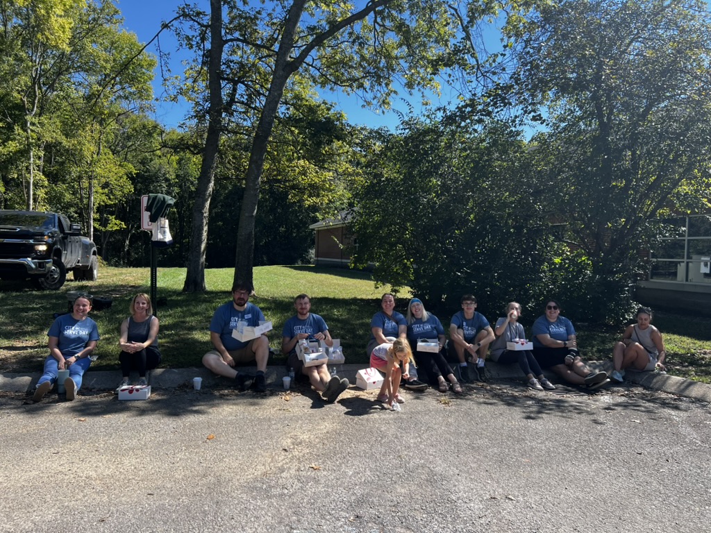 A group of people sit on a curb in a shaded area, enjoying food and drinks on a sunny day. Trees and houses are visible in the background, and a black truck is parked nearby.