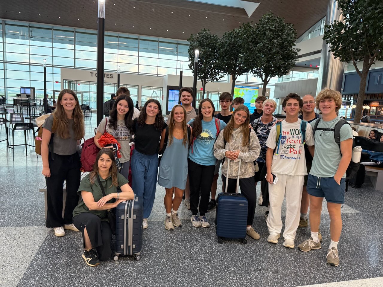A group of thirteen smiling young adults, some holding luggage, pose together in an airport terminal near seating and trees, ready for travel.