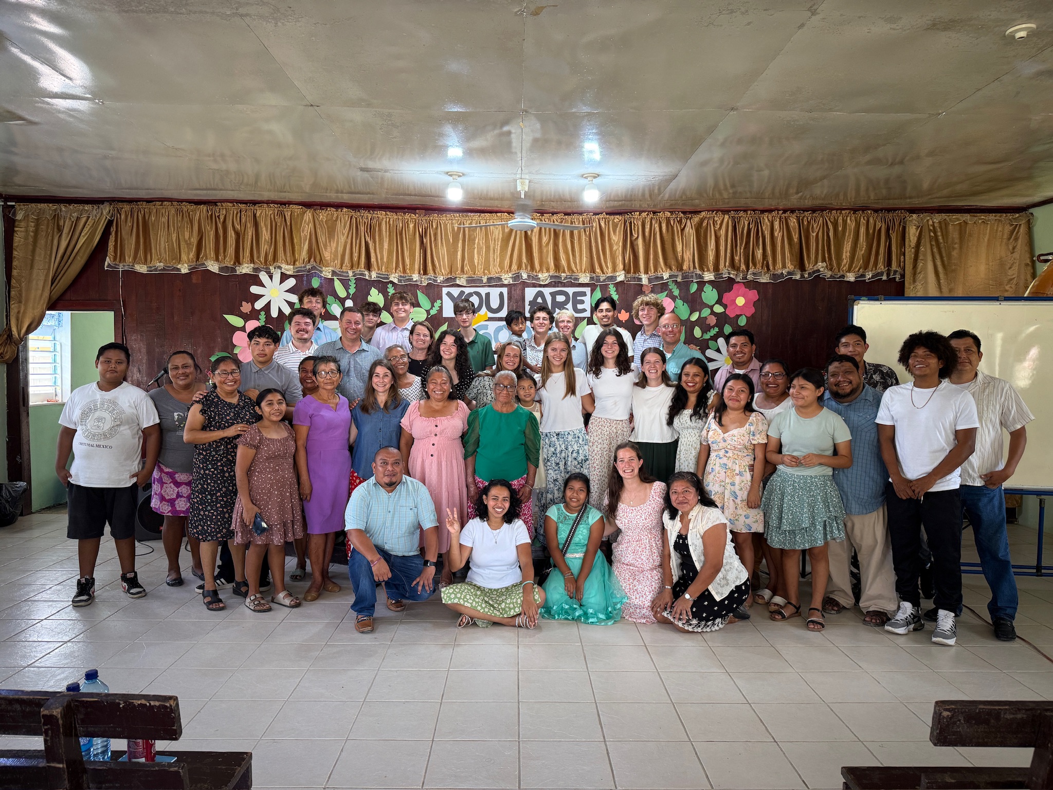 A large group of people of various ages pose together and smile inside a decorated room with a banner reading YOU ARE and colorful flowers on the wall behind them. Some are standing while others kneel or sit in front.