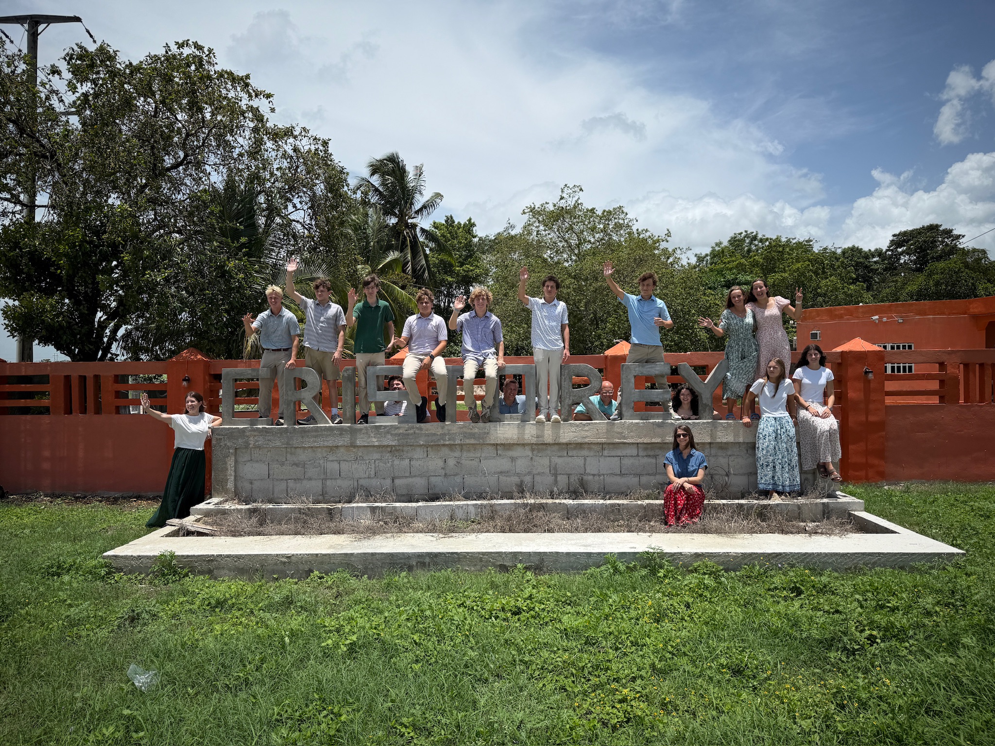 A group of people stands and sits on steps in front of large block letters spelling FELIPE CARRILLO PUERTO with a bright orange fence behind them, waving and smiling on a sunny day.