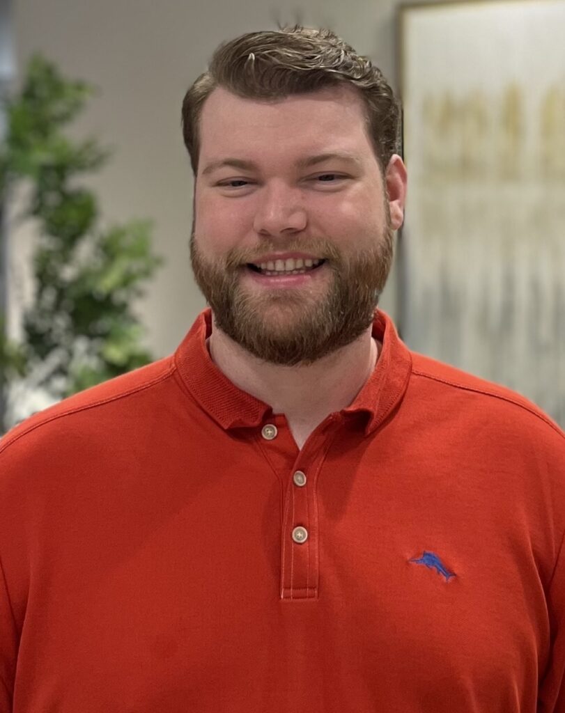 A man with short brown hair and a beard, wearing a bright red polo shirt, smiles while standing indoors. The background features blurred greenery and abstract wall art.