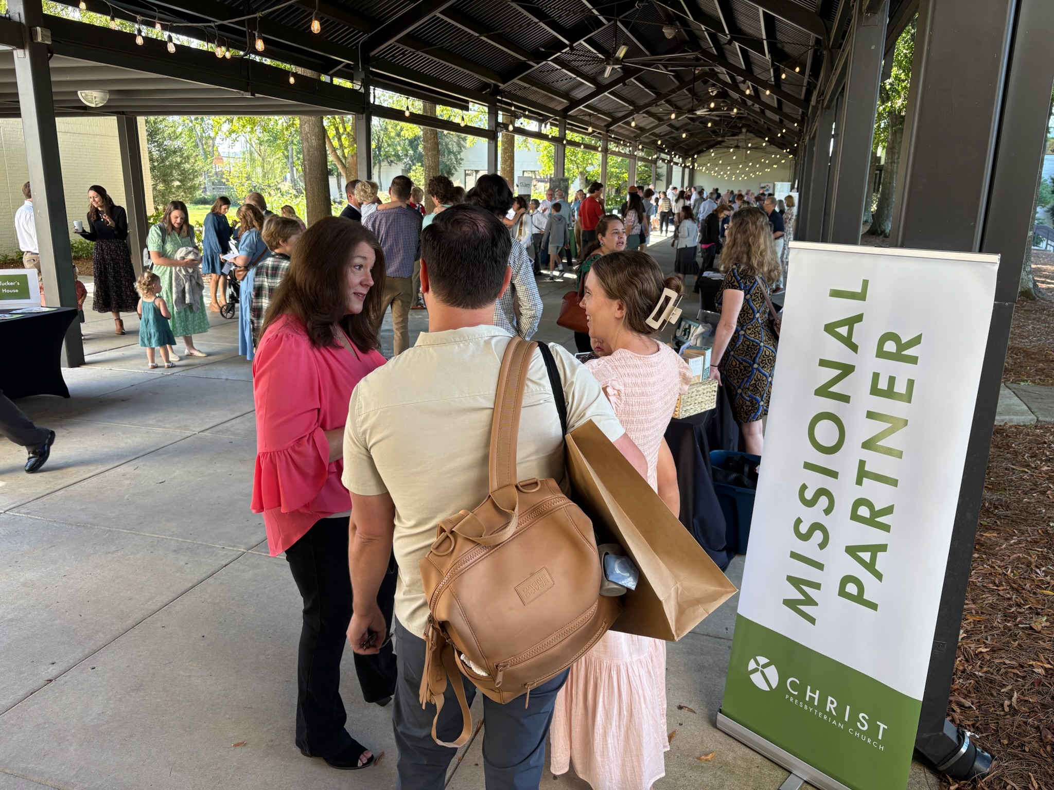 People gather and converse under a covered outdoor area at an event. A sign in the foreground reads Missional Partner with the Christ Covenant logo. Some attendees carry bags and wear casual clothing.