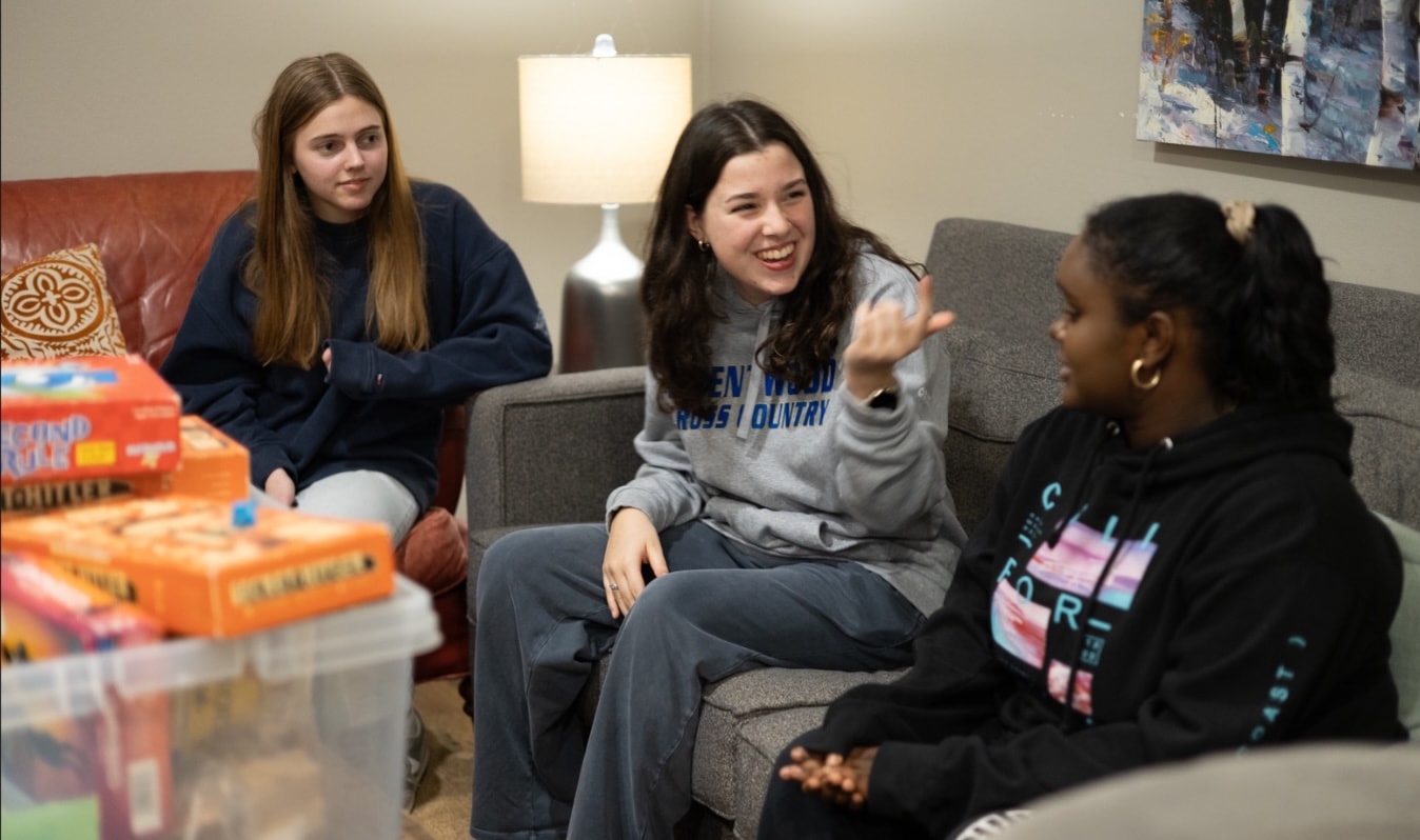 Three young women sit together indoors, talking and smiling. Board games are stacked on a table beside them. One woman gestures animatedly as the others listen. The setting appears cozy, with a lamp and artwork in the background.