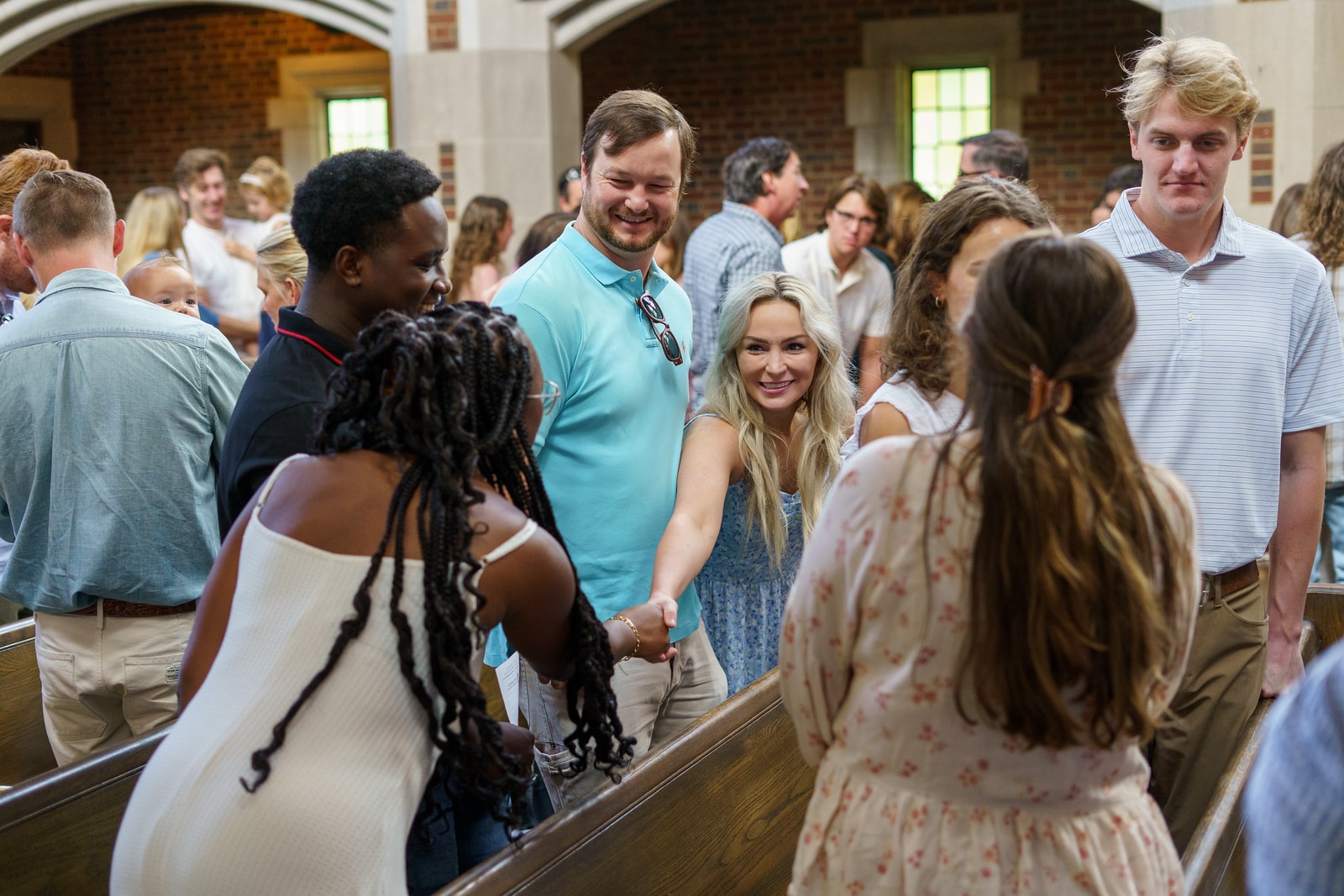A group of young adults stand and smile while greeting each other inside a church or chapel, some shaking hands, with sunlight coming through the windows in the background.