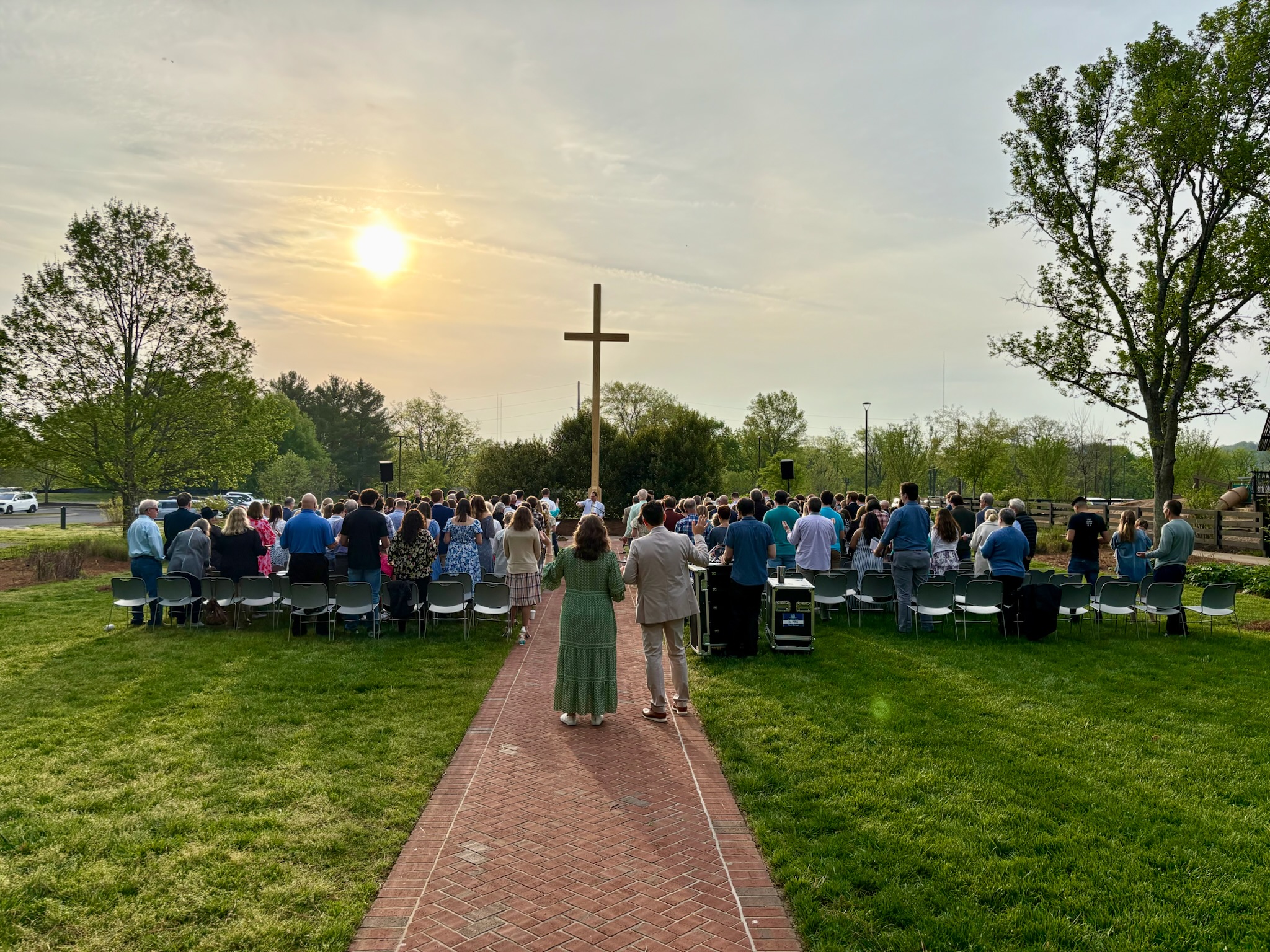A large group of people gather outdoors at sunrise, facing a wooden cross. They are seated on chairs arranged on a lawn, with trees and a clear sky in the background. Two people walk toward the group down a brick path.