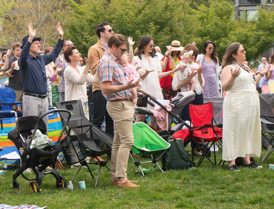 A group of people stand outdoors on a grassy area with folding chairs and blankets, raising their hands as if in celebration or worship. Trees and modern buildings are visible in the background.