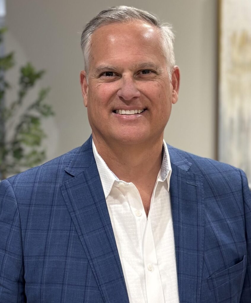 A middle-aged man with short gray hair, wearing a blue plaid suit jacket over a white dress shirt, smiles while standing indoors with a blurred background of greenery and artwork.