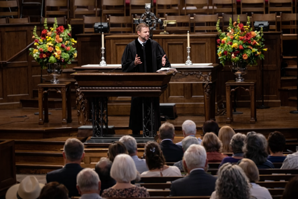 A man in a black robe speaks from a wooden pulpit to a seated audience in a church, with flower arrangements and candles on either side of the altar behind him.