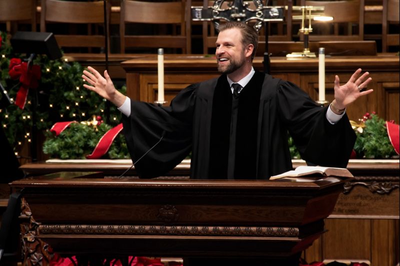 A man in a black robe stands at a wooden pulpit with open arms, smiling while speaking. Behind him are holiday wreaths, red ribbons, candles, and wooden chairs, suggesting a festive church setting.