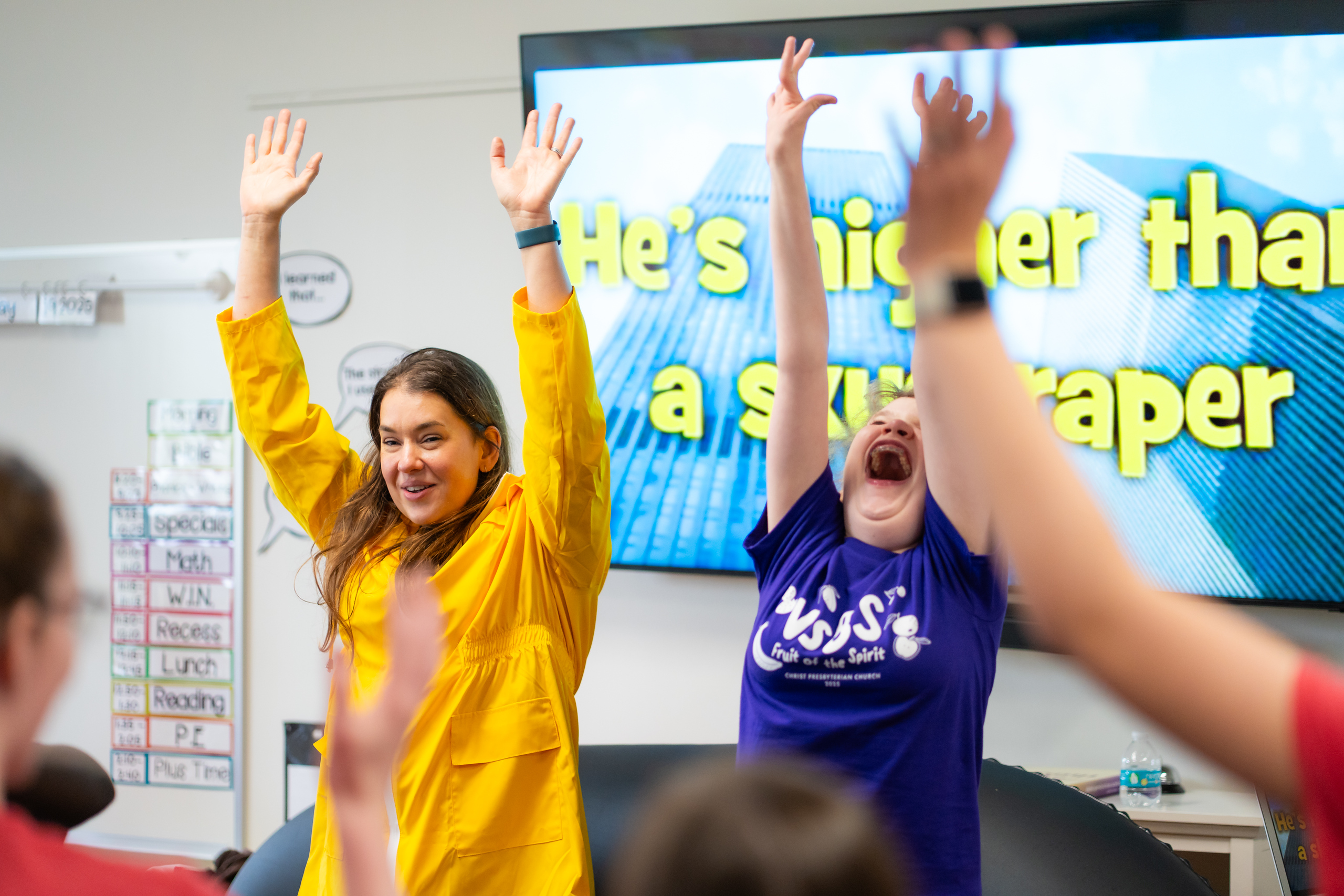 Two women with raised arms smile enthusiastically in a classroom, with children joining in. Behind them, a screen displays the words “He’s higher than a skyscraper” against an image of tall buildings.