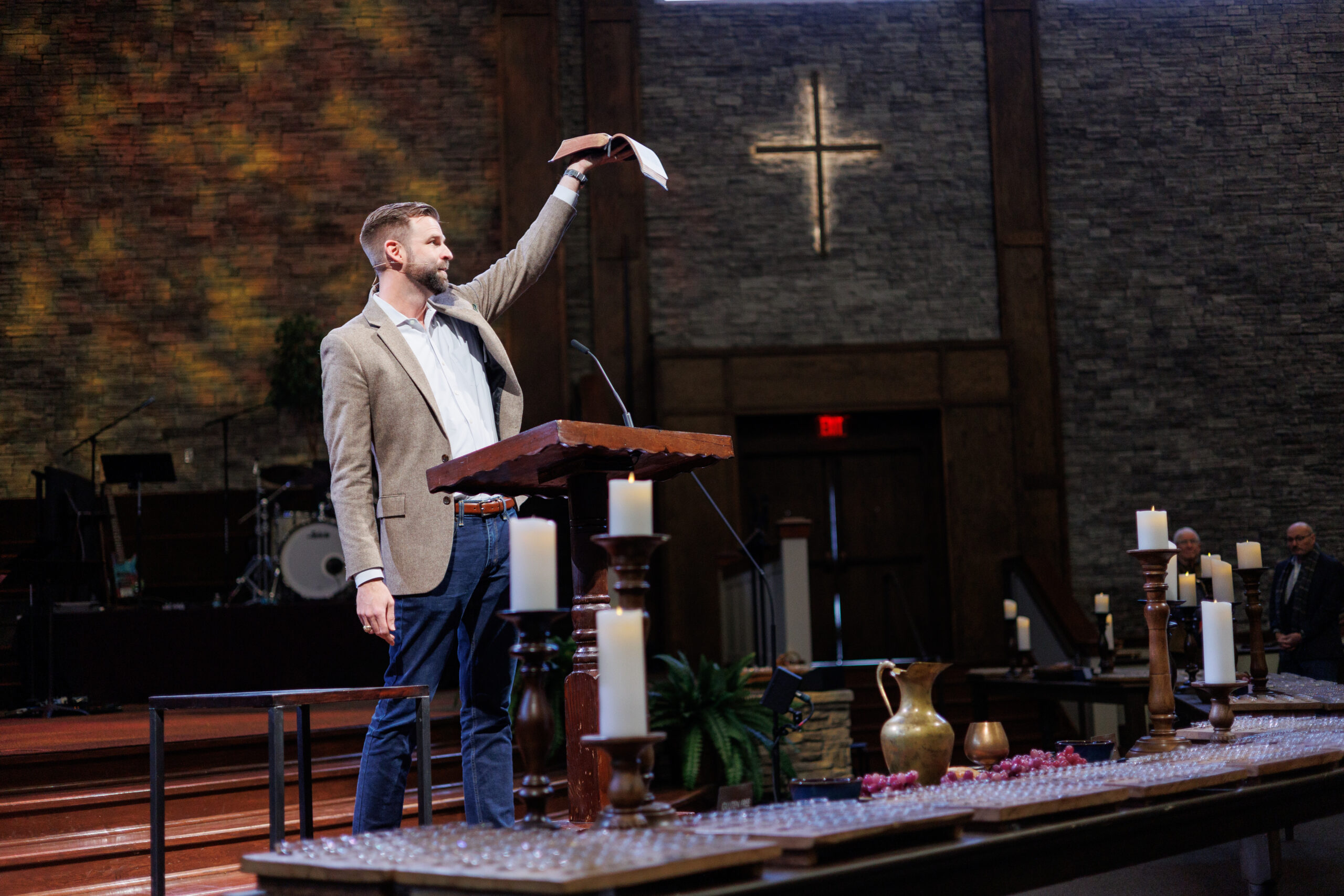 A man in a tan blazer stands at a wooden podium in a church, raising a paper in one hand. Candles and communion cups are arranged on tables in front of him, with a lit cross visible on the brick wall behind.