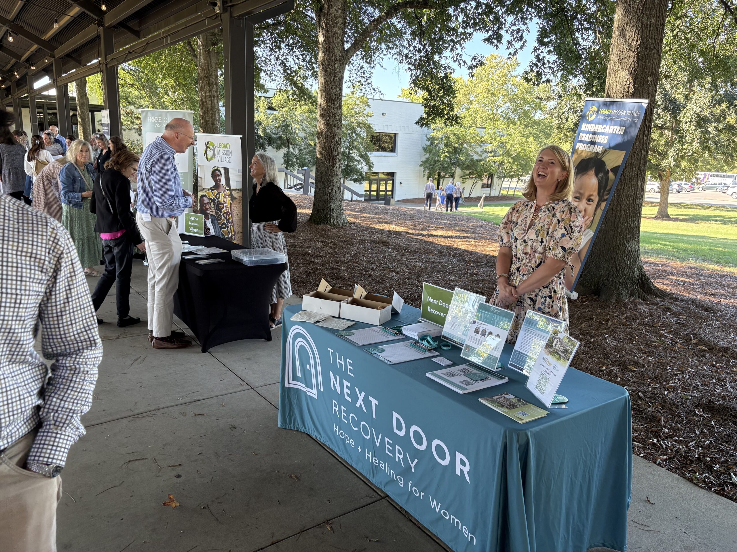 A woman smiles behind a table with The Next Door Recovery banner at an outdoor event, with brochures and signs on the table. Other booths and people are visible under a pavilion with trees and buildings in the background.