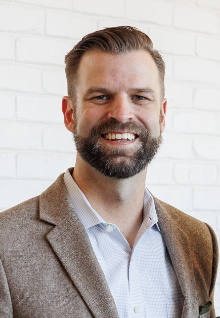 A man with short brown hair and a beard, wearing a brown blazer and a light-colored shirt, smiles in front of a white brick wall.