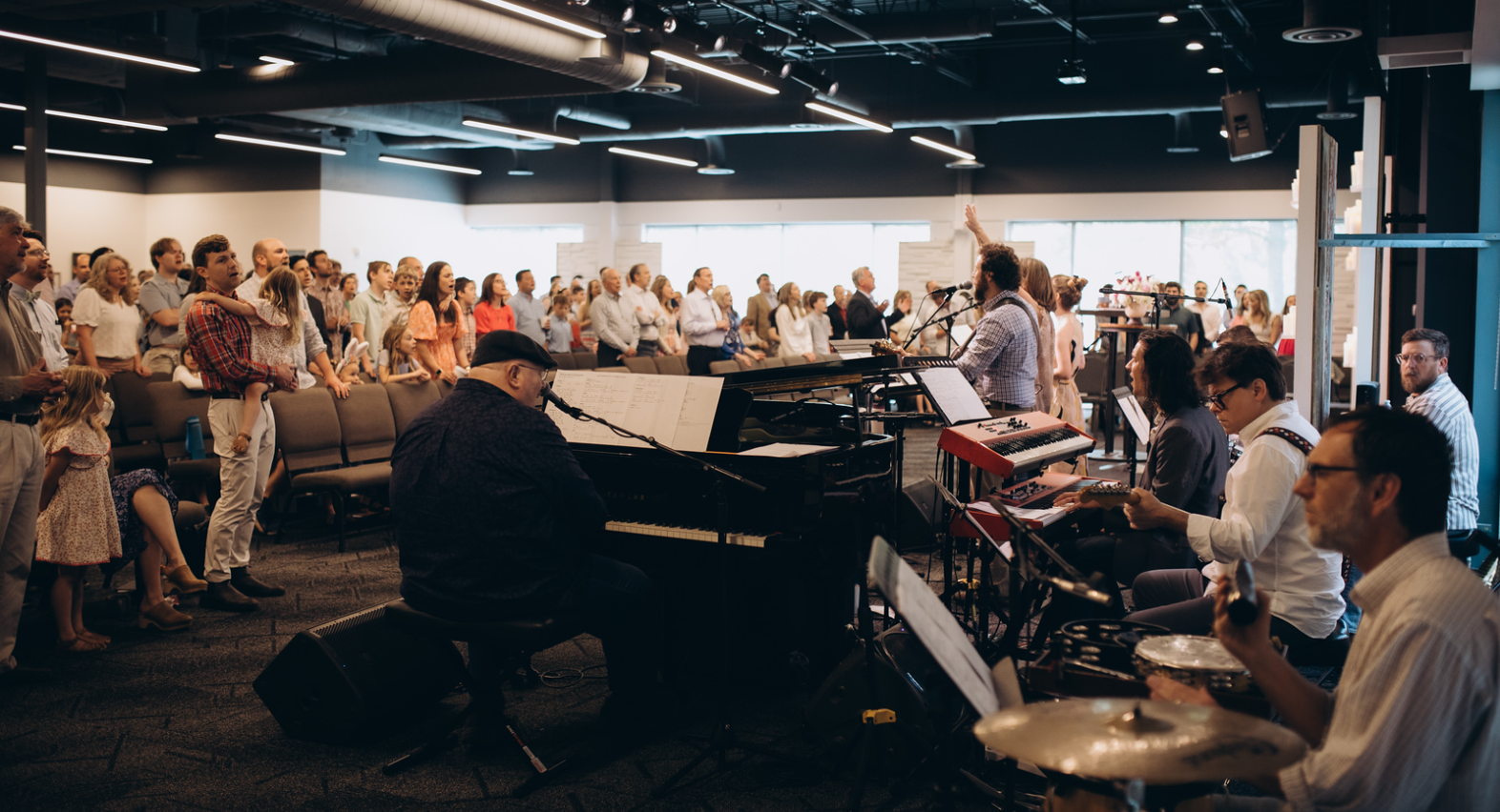 A band performs on stage with piano, drums, and guitars as a large congregation stands and sings along in a modern, well-lit church or gathering space.