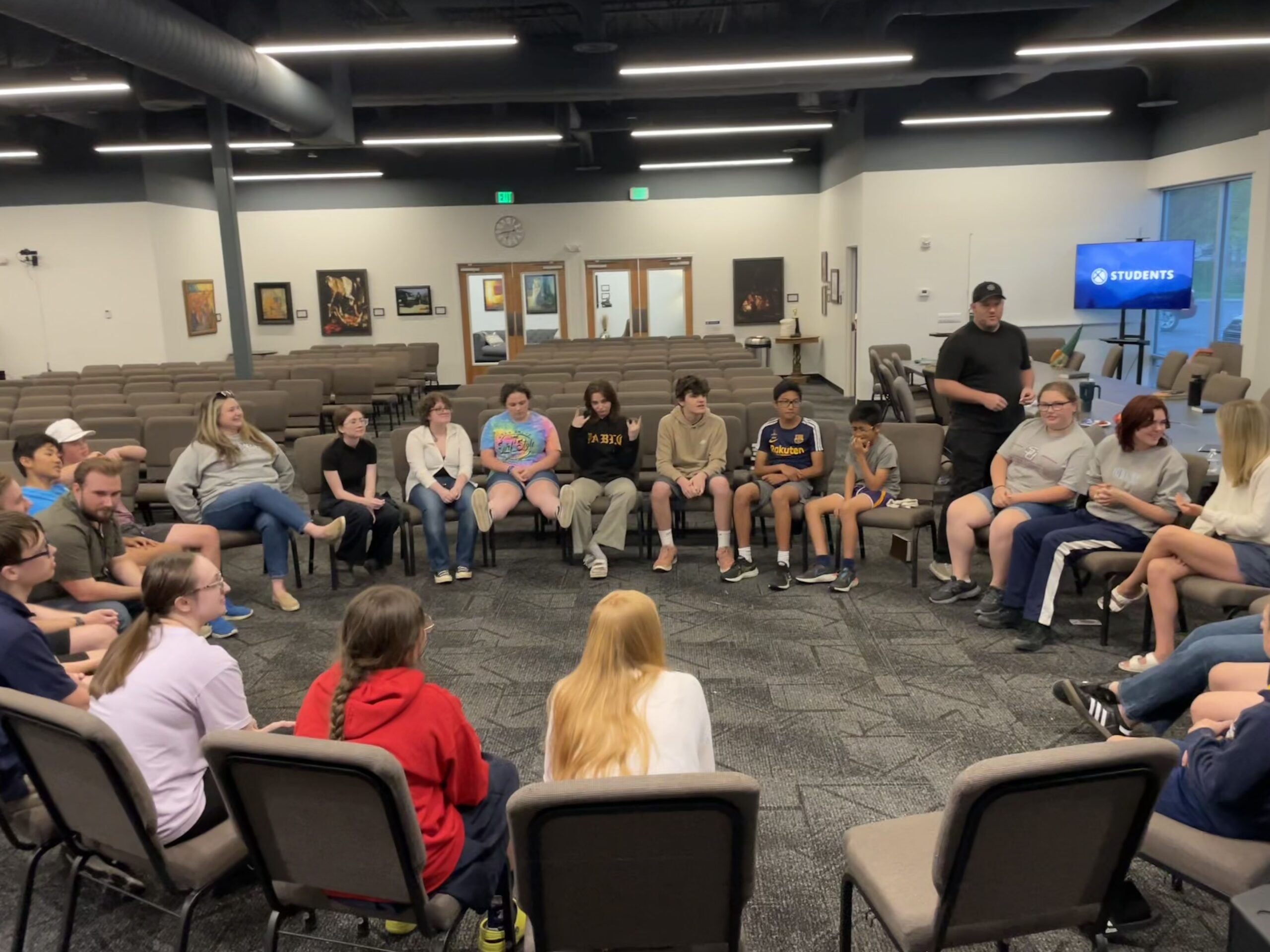 A group of teenagers and adults sit in a large circle of chairs inside a spacious room, engaged in conversation or activity, with a TV screen in the background displaying the word “STUDENTS.”.