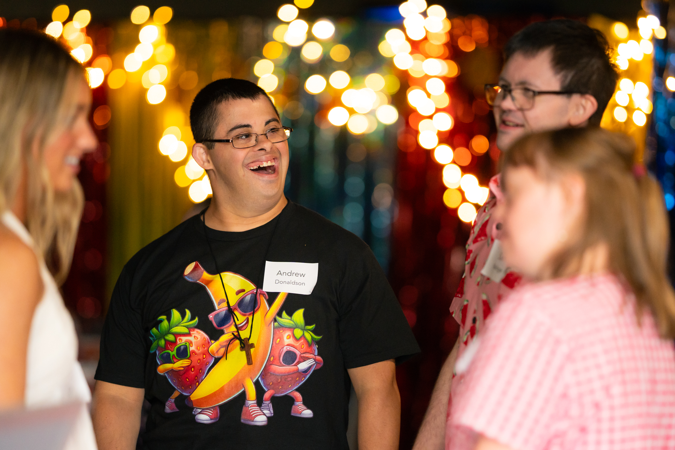 A young man wearing glasses and a fun t-shirt featuring cartoon fruits smiles while talking with three other people at a festive event with colorful lights in the background.