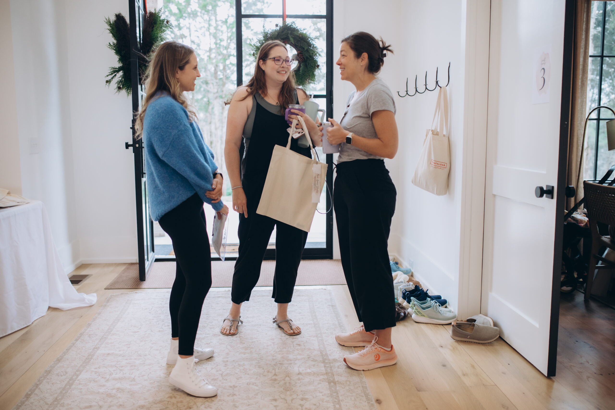 Three women stand and chat in a bright, modern entryway with a glass door. One holds a large tote bag, and another has a cup. Shoes and bags are scattered on the floor and hooks by the wall.