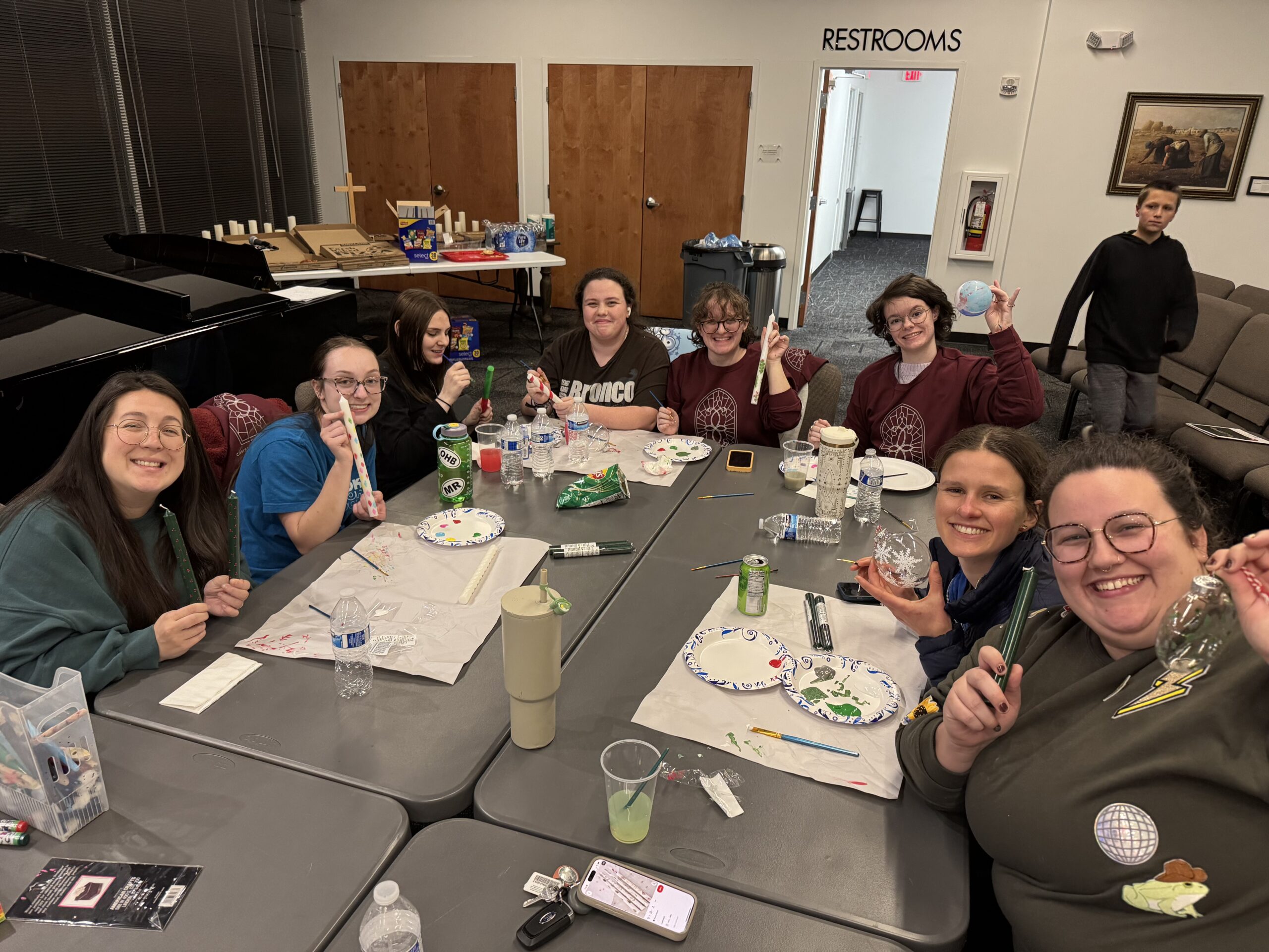 A group of nine smiling people sits around a table painting and holding up their art in a brightly lit room. Art supplies, drinks, and snacks are scattered on the tables. A young boy stands in the background near the “restrooms” sign.
