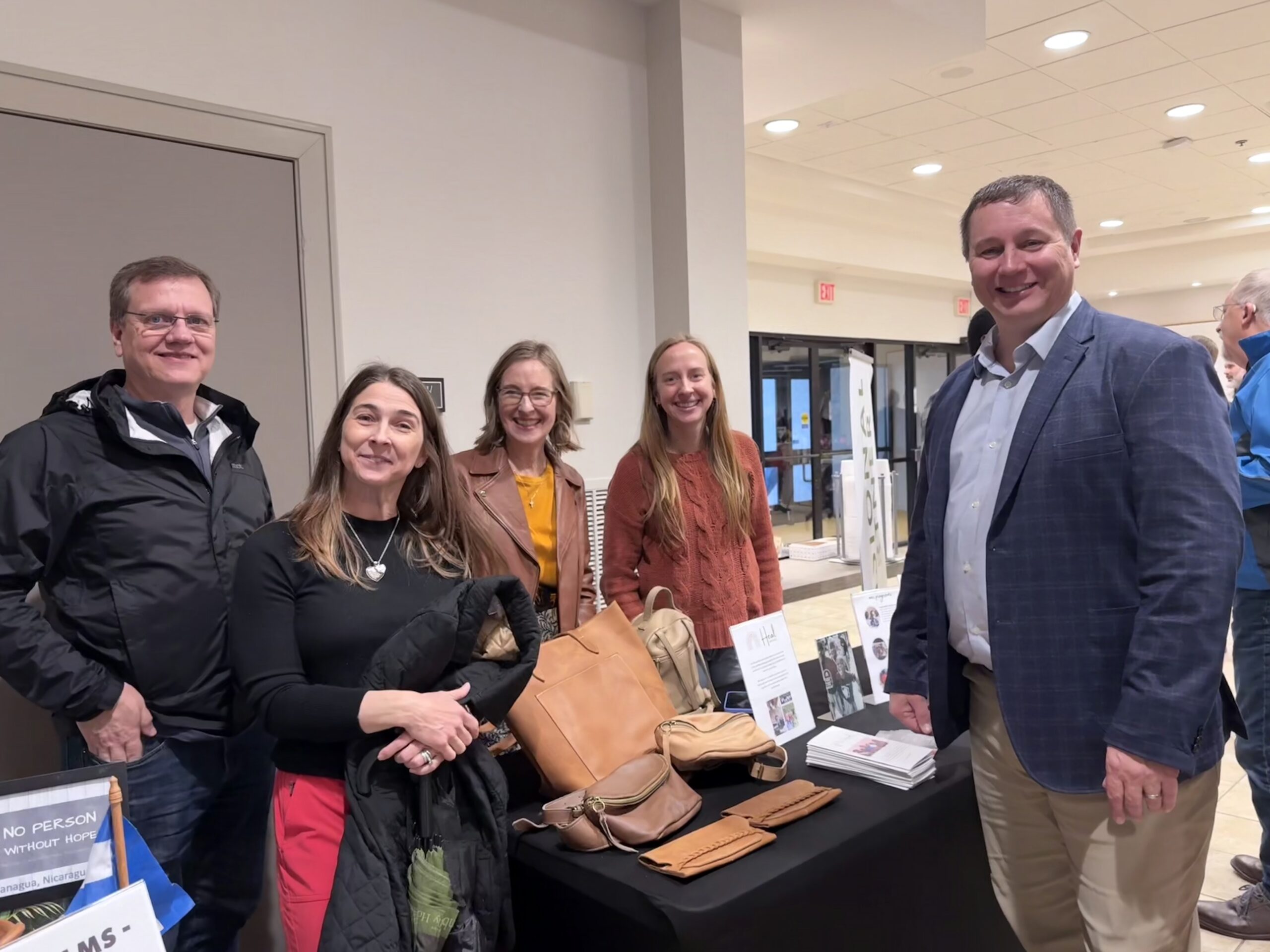 Five adults stand and smile around a table displaying books, flyers, and bags in a brightly lit indoor setting with white walls and ceiling lights.