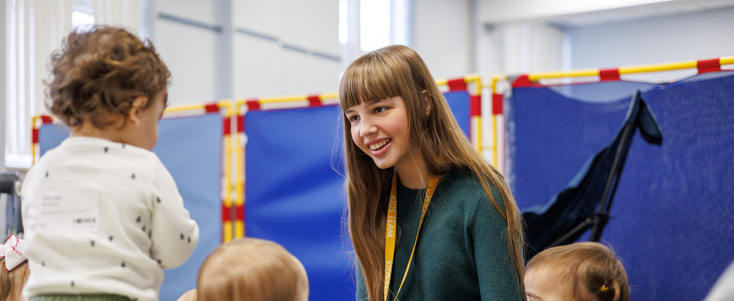 A young woman sits on the floor, smiling and engaging with a group of toddlers in a brightly lit indoor play area with colorful barriers in the background.