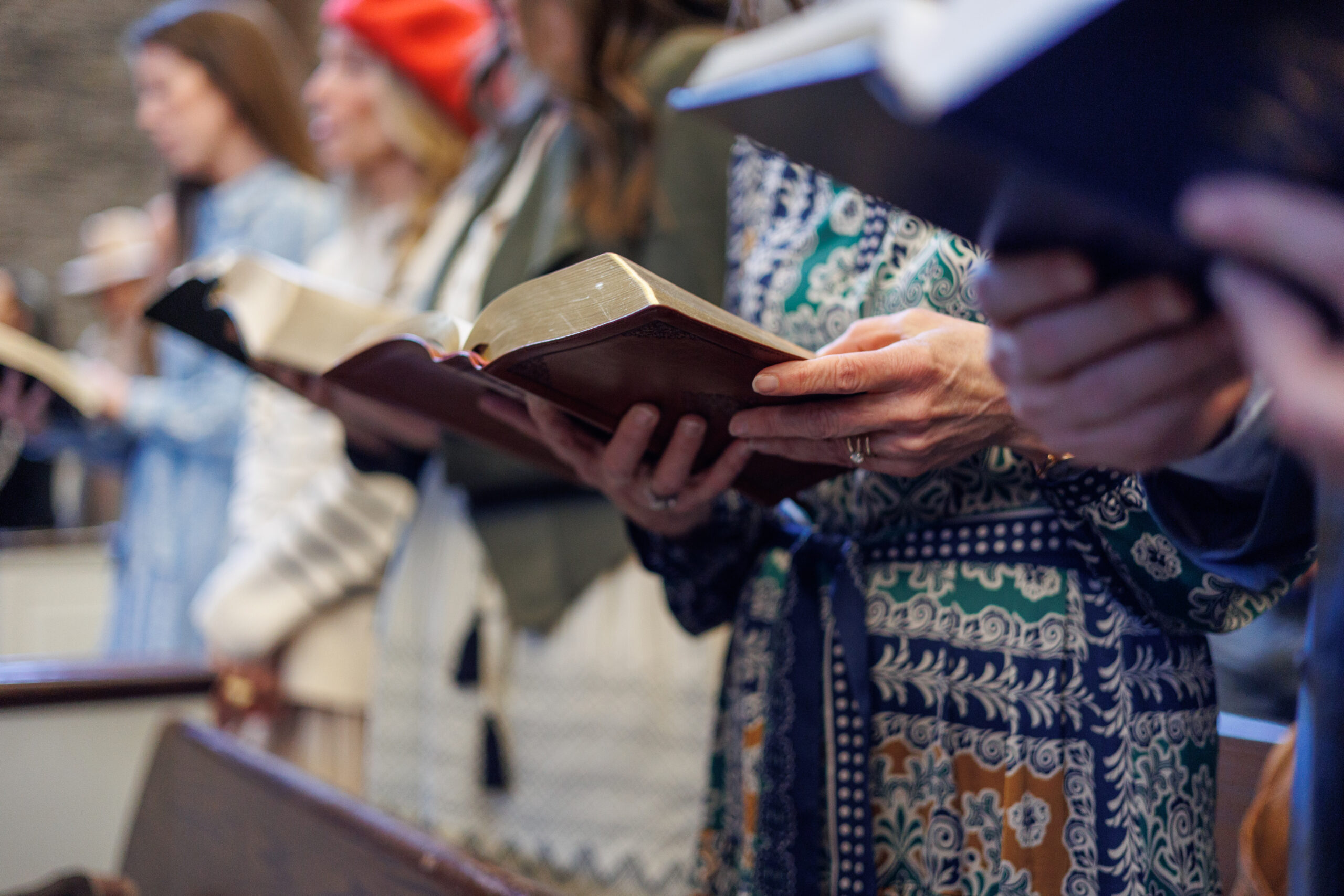 A group of people standing in church pews, holding open hymnals or books, with their faces out of focus. They appear to be singing or participating in a religious service.