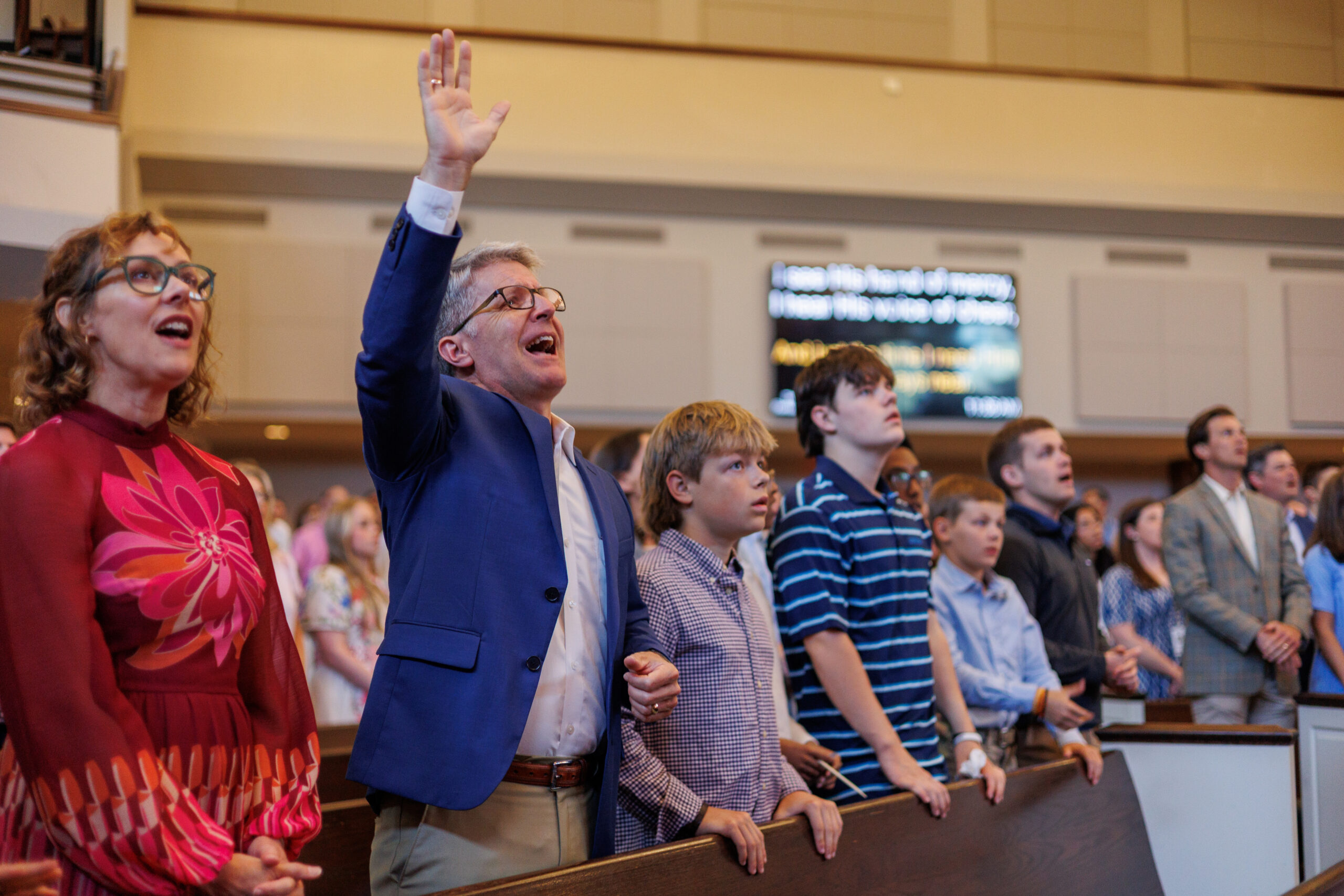 A group of people stand in church pews during a service; a man in a blue blazer raises his hand while others sing or watch attentively. Children and adults are dressed in formal attire.