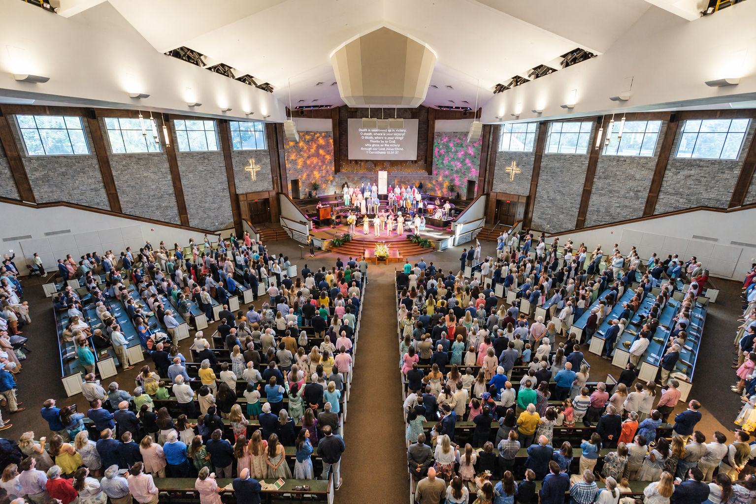 A large congregation gathers in a spacious, modern church sanctuary for a service, with people seated in rows and a choir and clergy standing at the front near colorful decorations and a cross.