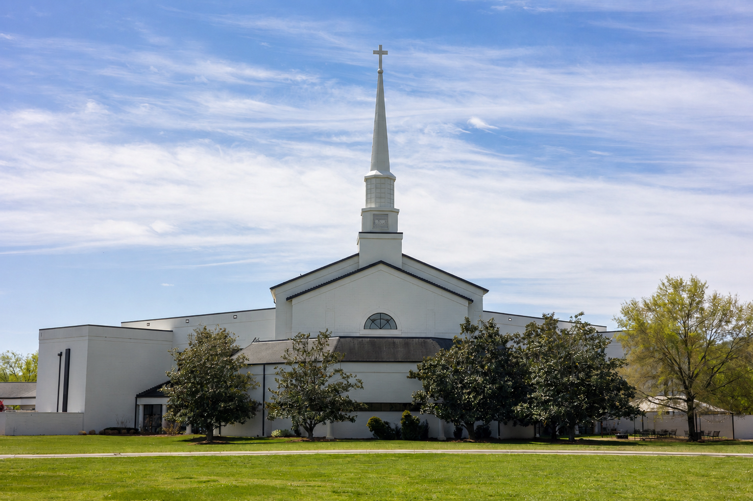 A white church with a tall steeple topped by a cross stands under a blue sky with wispy clouds, surrounded by green grass and trees.