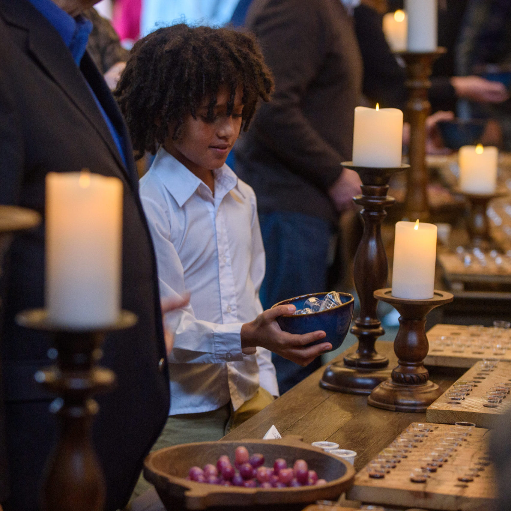 A young boy in a white shirt holds a bowl at a candlelit table filled with small items and surrounded by people, suggesting a ceremonial or festive gathering.