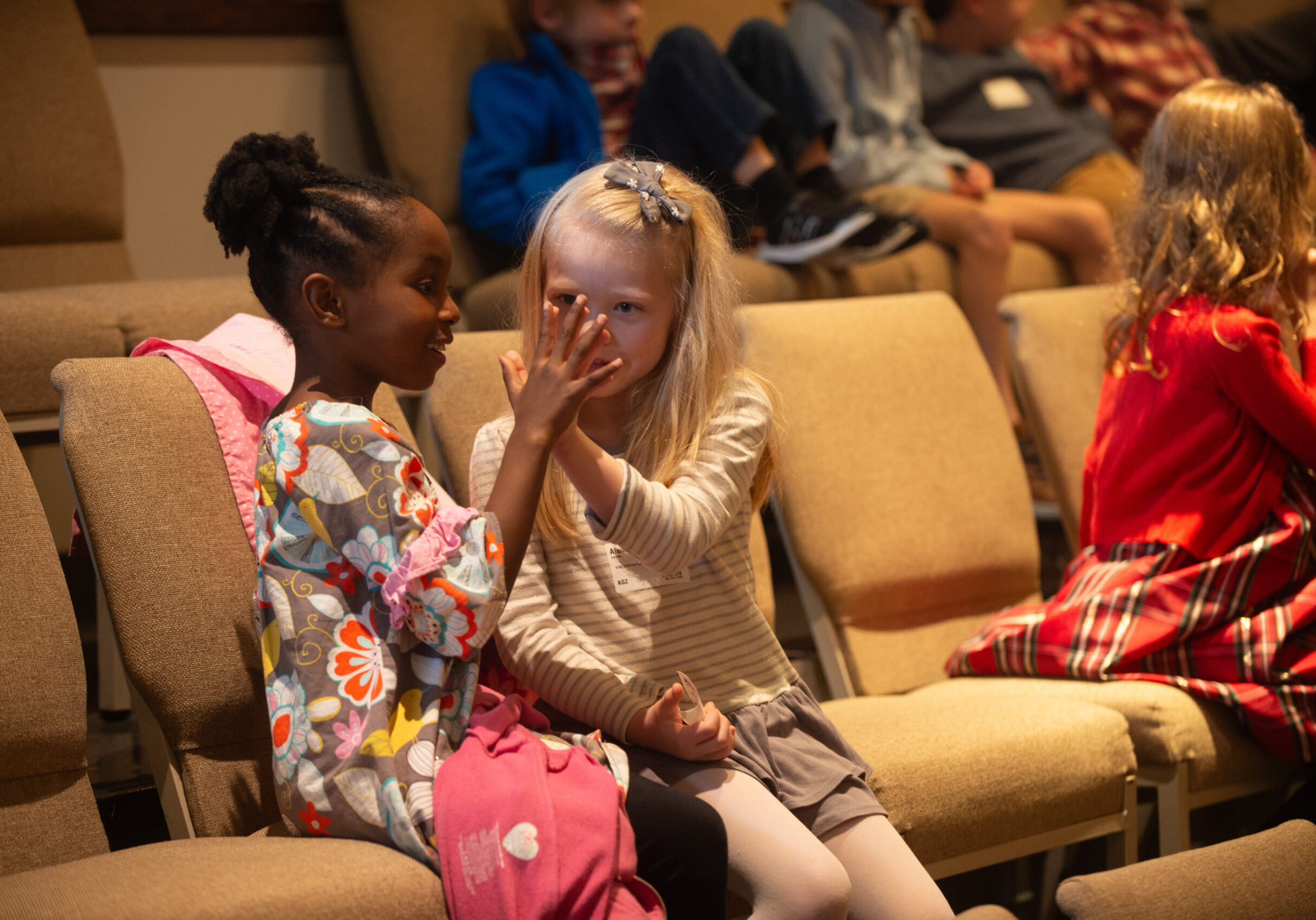 Two young girls sit close together on beige chairs, one with her hand raised as if showing something to the other. They appear to be engaged in a friendly conversation. Other children are seated in the background.
