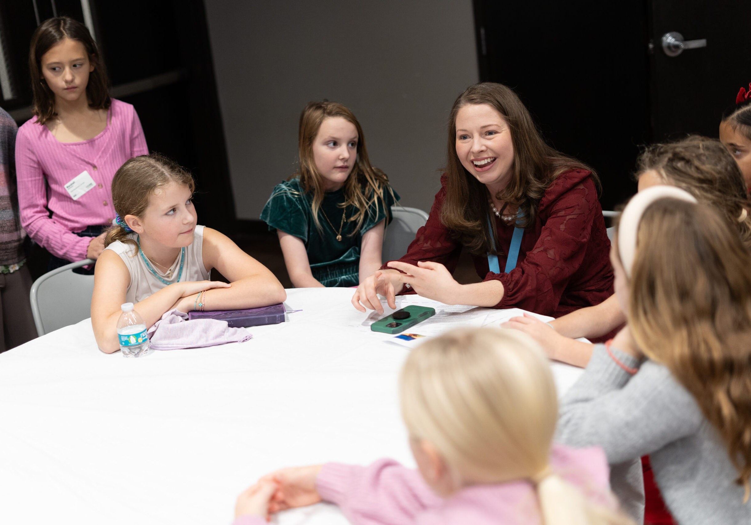 A group of young girls sits around a table, listening to a smiling woman who appears to be leading a discussion or activity. The atmosphere is bright and engaging.