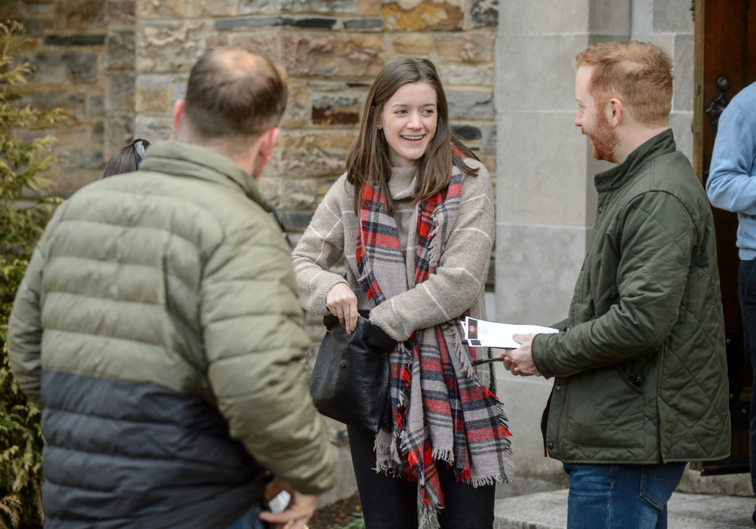 Three people stand and converse outside a stone building. A woman in a scarf smiles while holding her bag, and a man in a green jacket holds a pamphlet. Another person faces away from the camera.