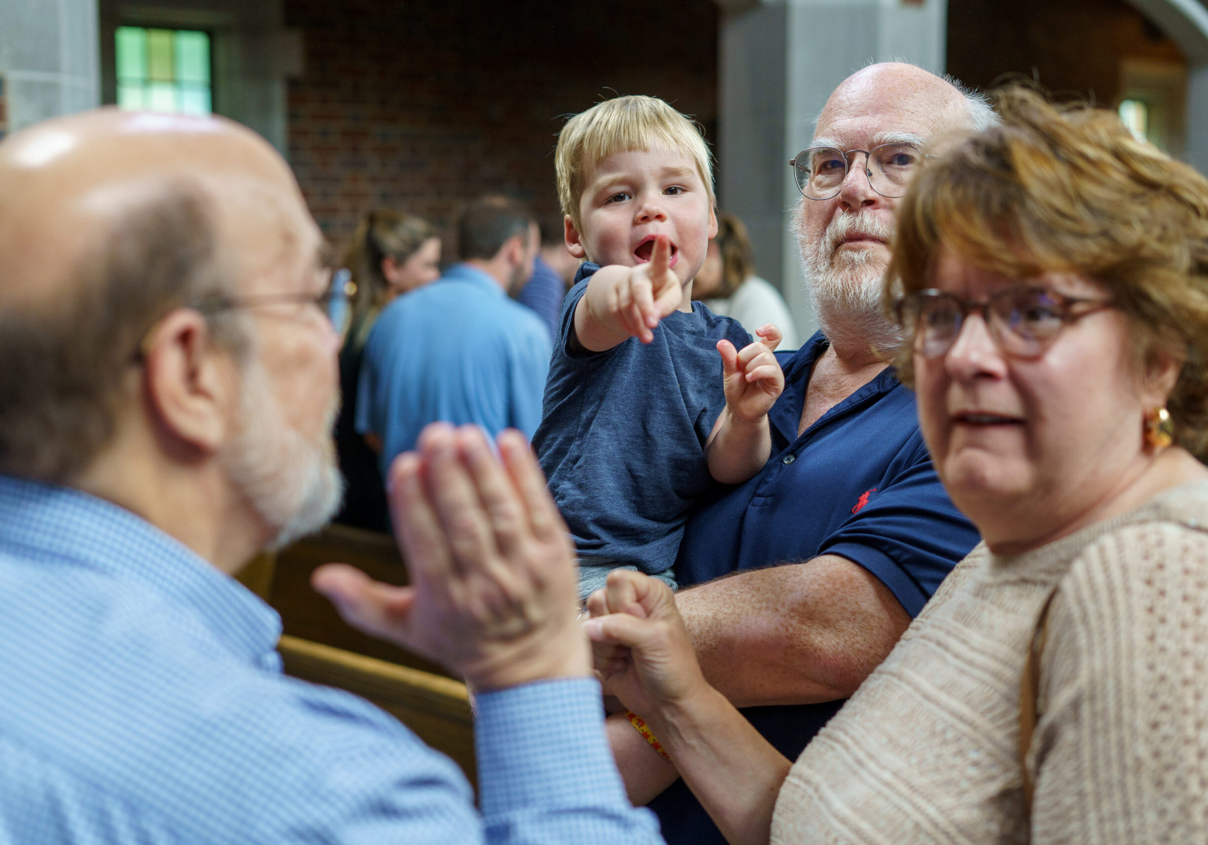 A young boy points toward the camera while being held by an older man in a church setting. Two other adults stand nearby, all looking in the same direction. The background shows pews and other people.