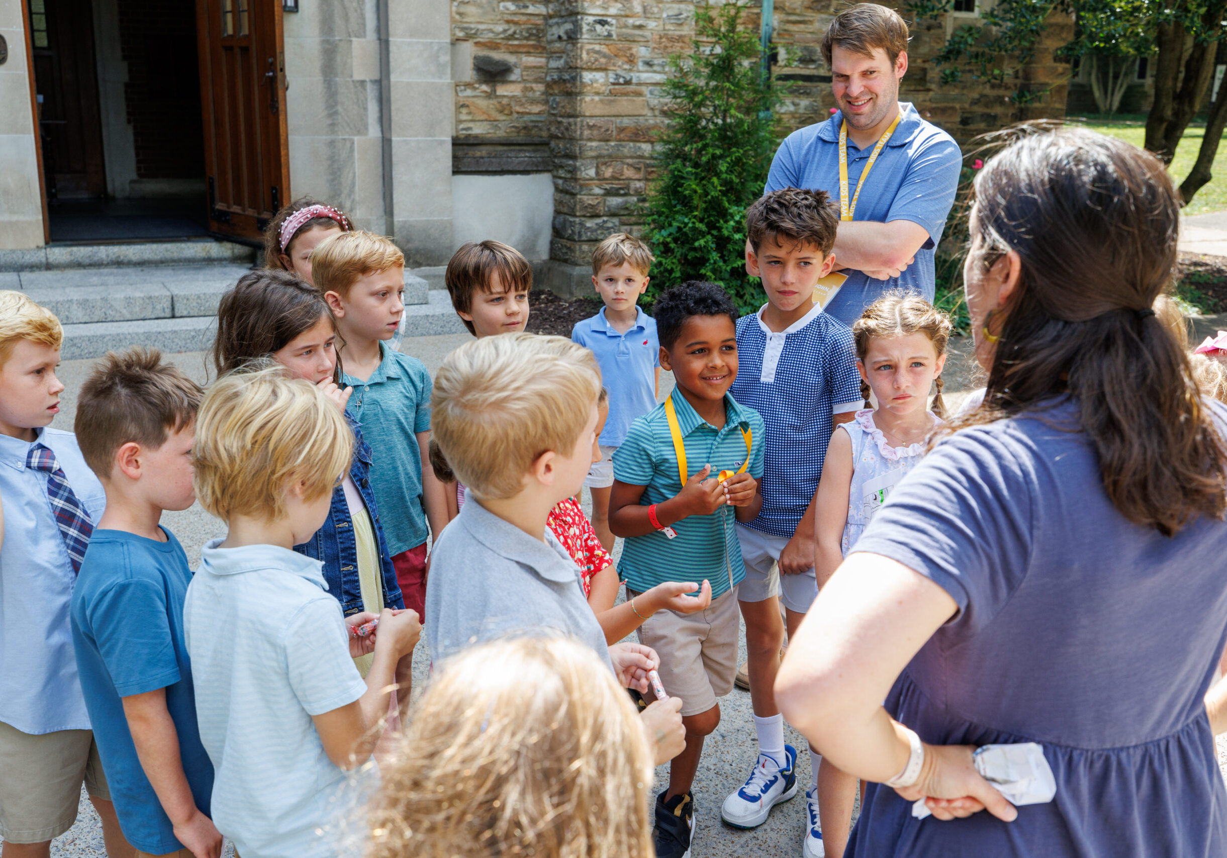 A group of young children stand outside a stone building, gathered around two adults who appear to be giving instructions. The children look attentive, some wearing casual clothes, while the adults wear lanyards.