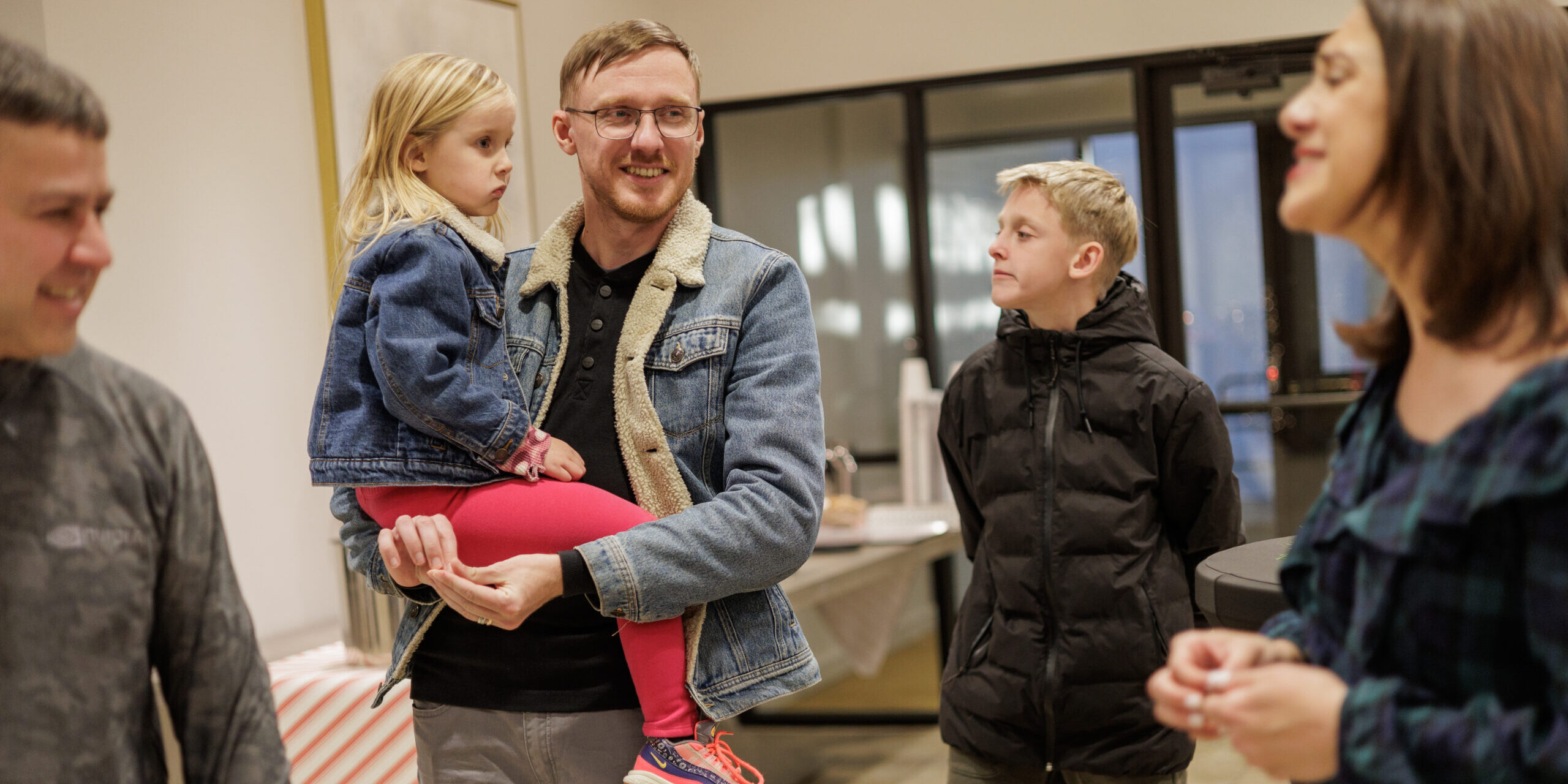 A man holding a young girl stands smiling with three other people in a casual indoor setting. The group appears to be talking and enjoying each others company.