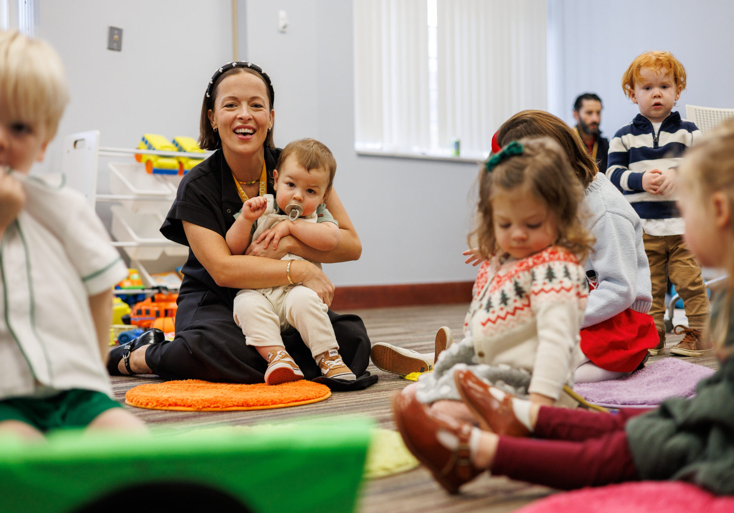A smiling woman sits on a colorful mat holding a baby with a pacifier, surrounded by young children sitting and playing in a bright classroom. Toys are visible in the background.