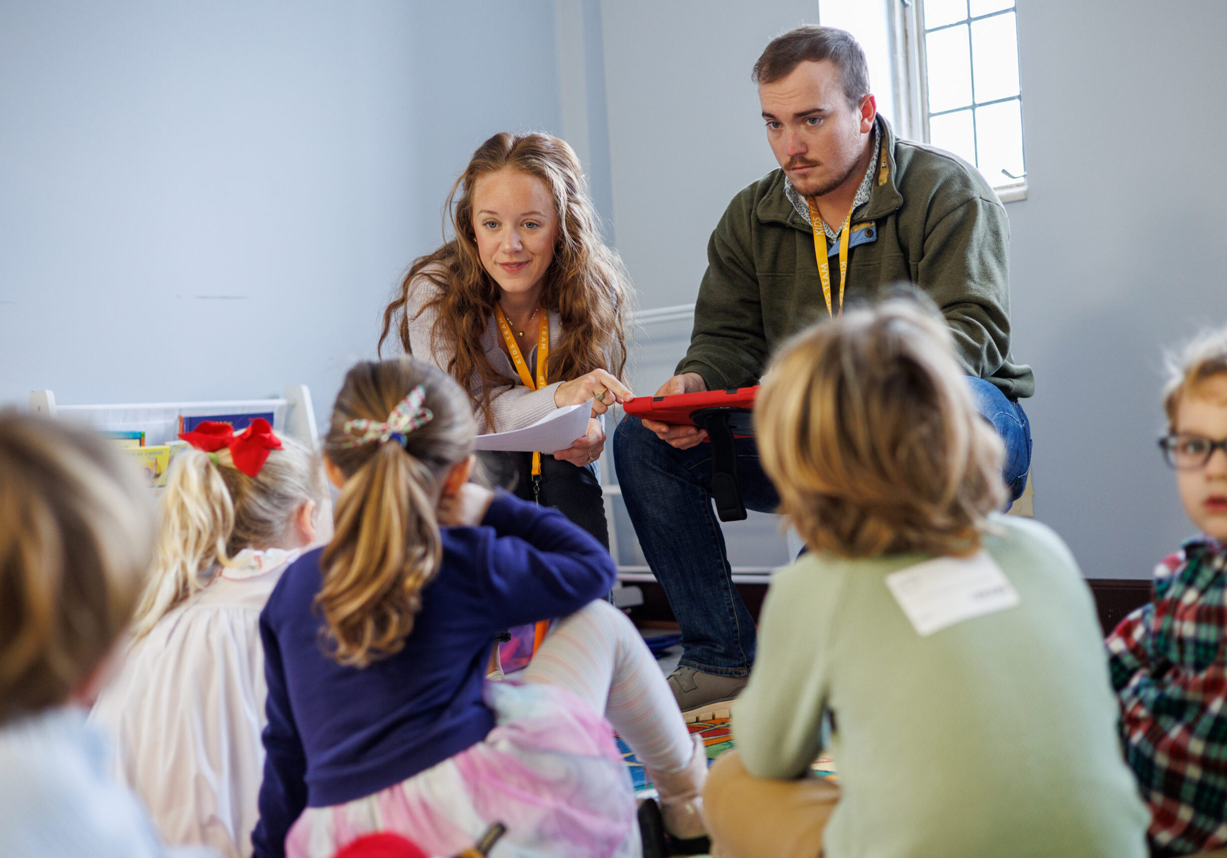 Two adults sit on the floor, engaging with a group of young children who are sitting and facing them in a classroom setting. The adults appear to be leading an activity or discussion.