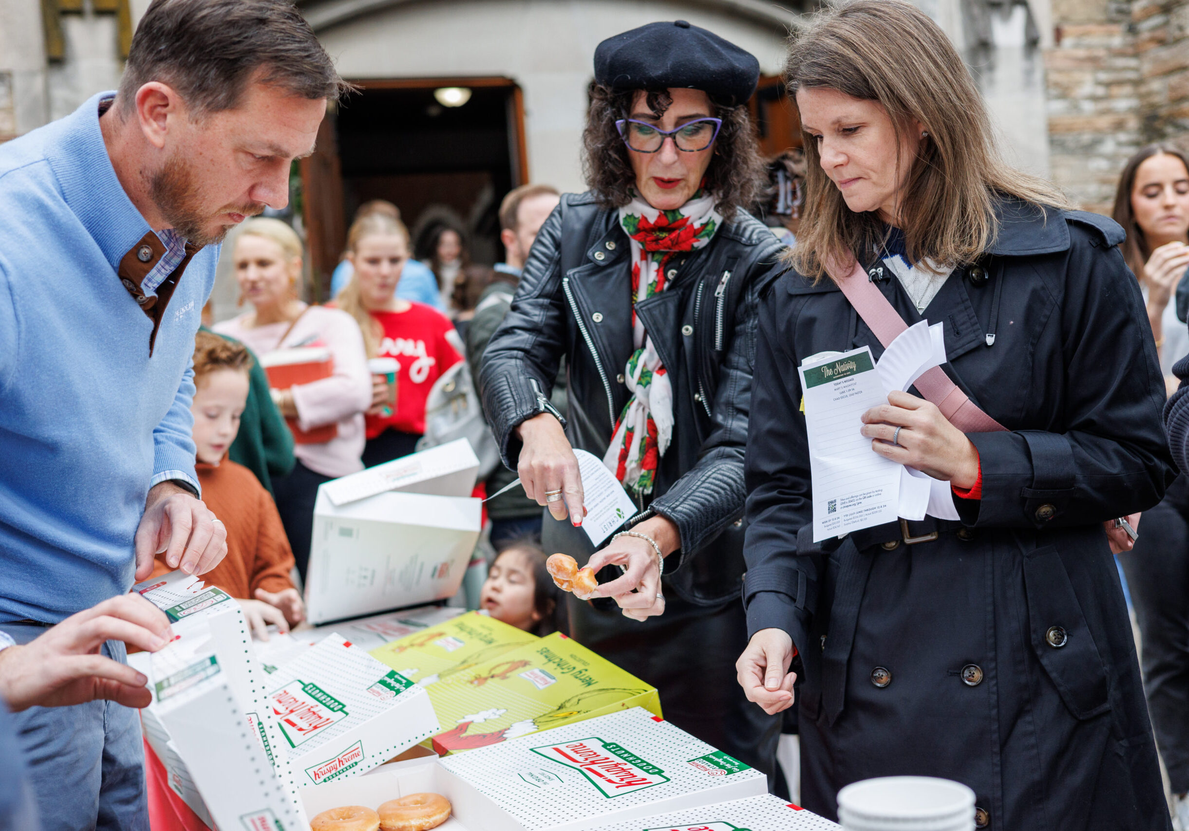 A group of people gather around a table with Krispy Kreme donut boxes outdoors; two women are selecting donuts while others wait in line behind them.