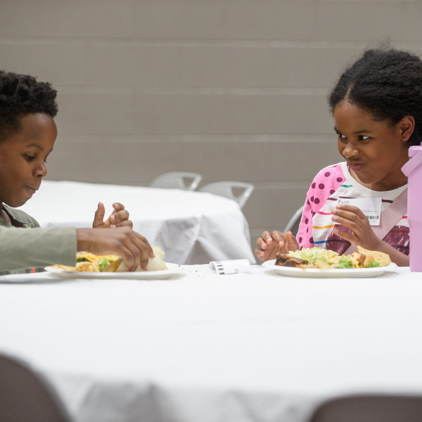Two children sit at a round table with white tablecloths, eating a meal together. The girl in a pink shirt looks at the boy, who is focused on his food. Both have plates of food and a purple water bottle is nearby.