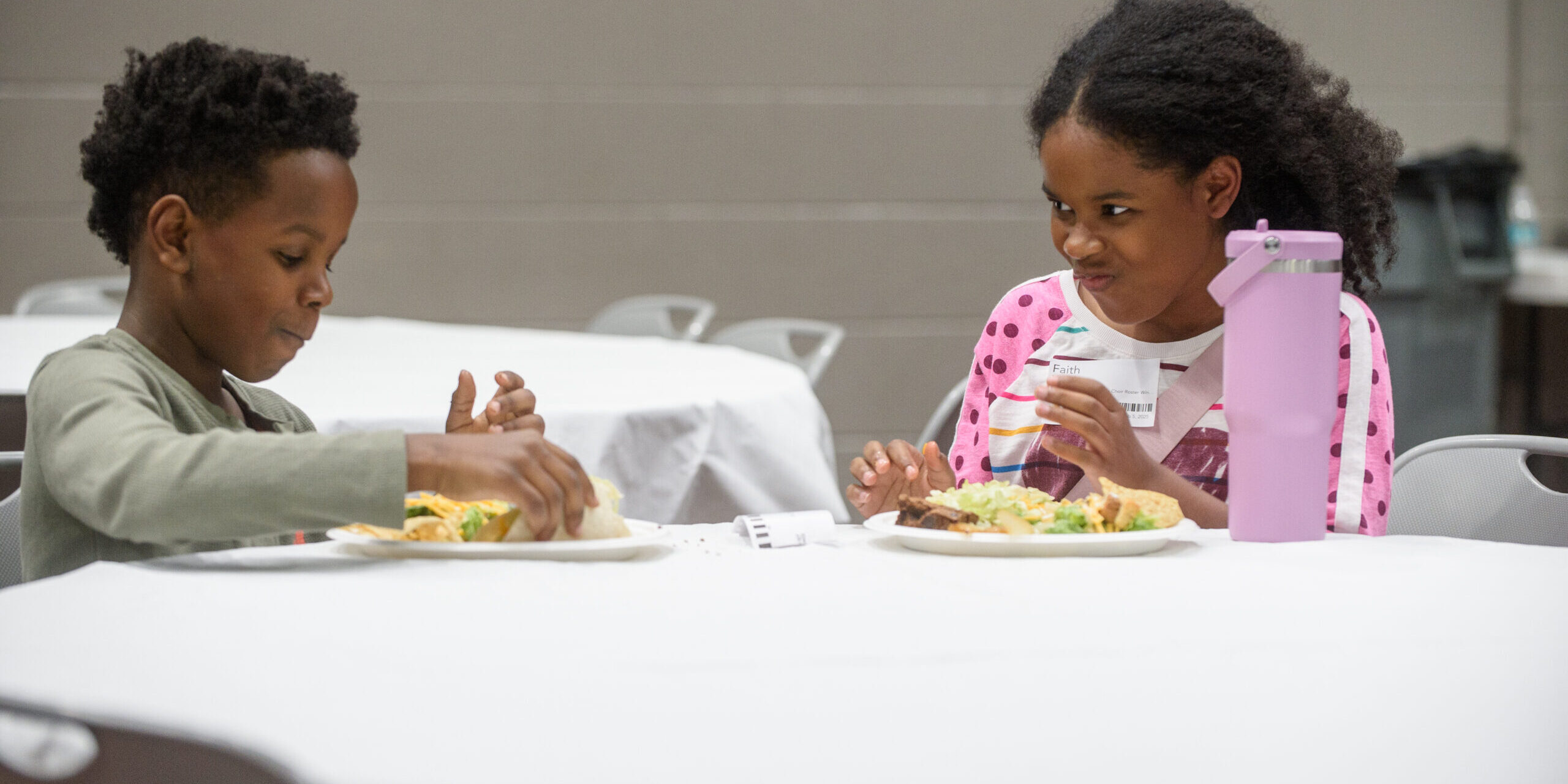 Two children sit at a round table eating a meal together. The girl in a pink polka dot sweater smiles and looks at the boy, who is focused on his food. A large purple water bottle is on the table.